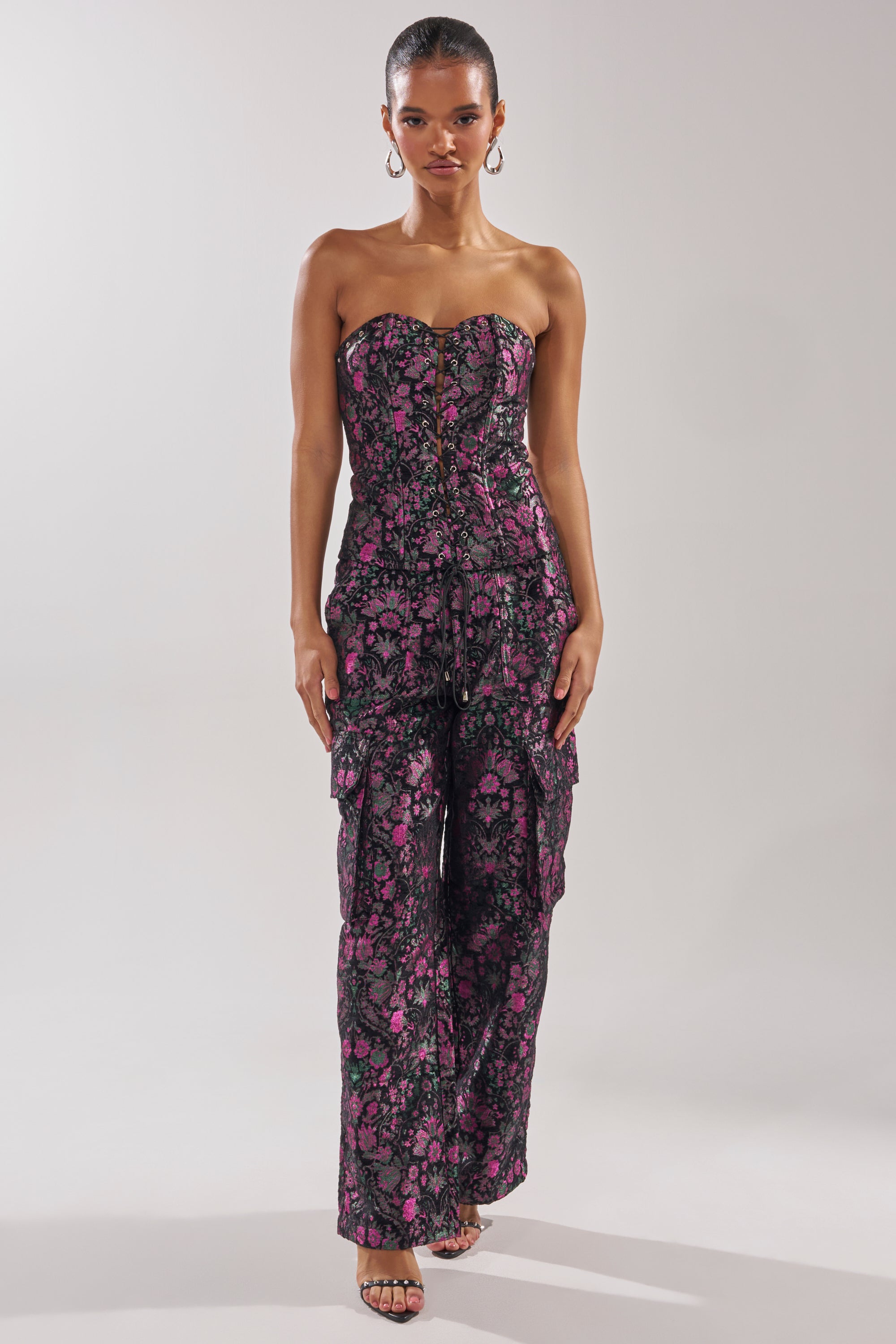 A woman stands against a plain background wearing the KIMMY FLORAL CORSET, a strapless, form-fitting black jumpsuit with metallic floral accents and ruffled layers, paired with strappy heels and hoop earrings.