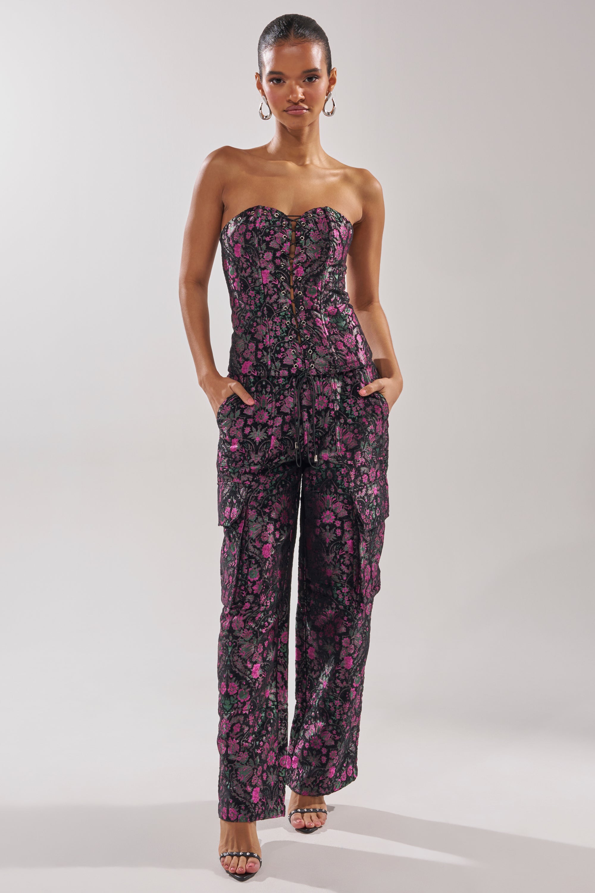 A woman stands confidently in the KIMMY FLORAL CORSET, a strapless, fitted jumpsuit with a black background and pink floral pattern, featuring pockets. She accessorizes with large hoop earrings and open-toed heels.