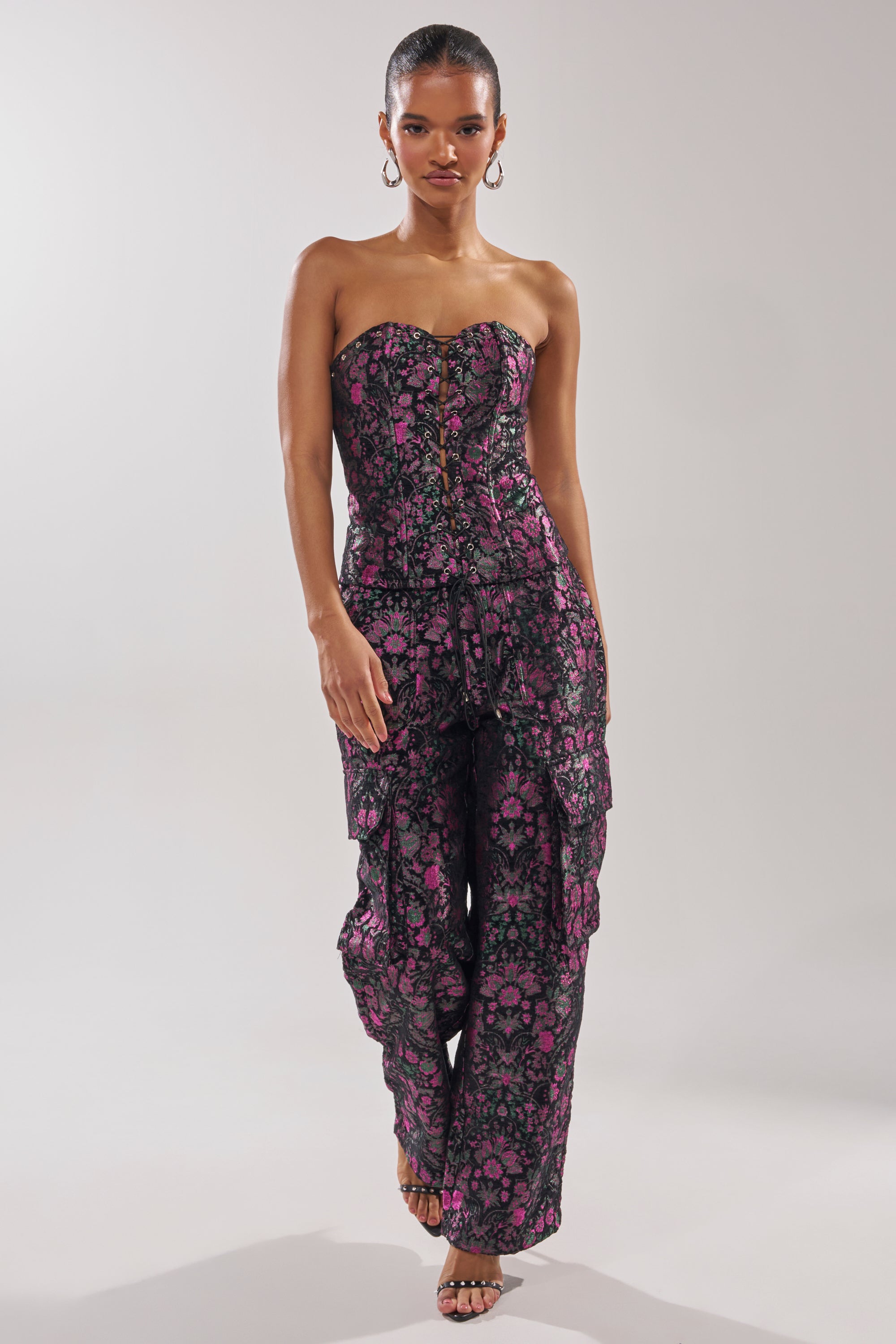 A woman confidently wears the KIMMY FLORAL CORSET, a strapless fitted jumpsuit with a black base, pink florals, and metallic detailing. She accessorizes with hoop earrings and strappy heels, set against a plain light gray background.