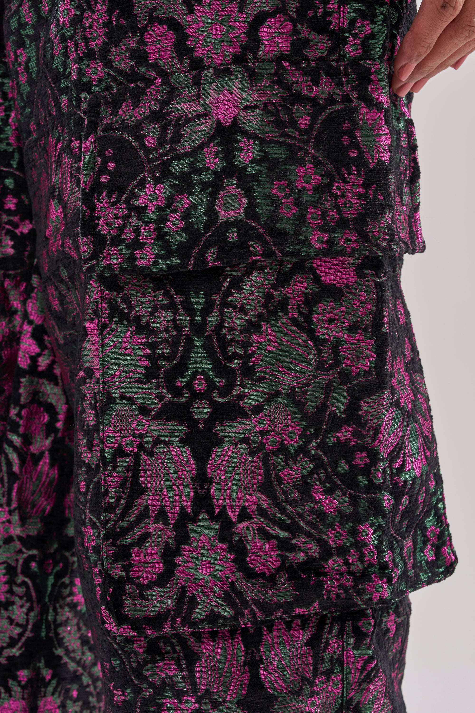 Detailed close-up of fabric with ornate purple, magenta, and green floral patterns on black—similar to the KIMMY FLORAL TROUSER. A hand appears near the top right corner. 9