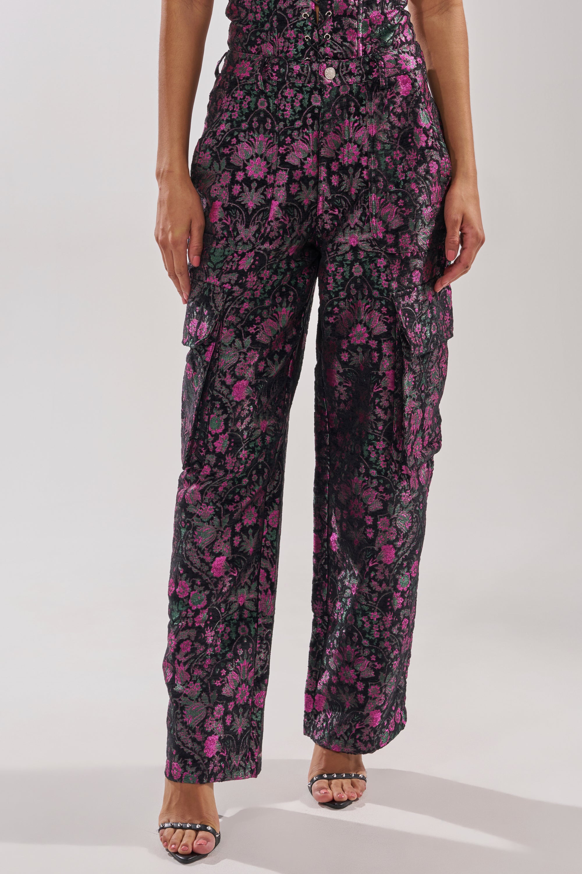 A person models the KIMMY FLORAL TROUSER—high-waisted cargo pants with a black base, vibrant pink blooms, and metallic floral accents—paired with open-toe heels in a cropped shoulders-down shot. 1