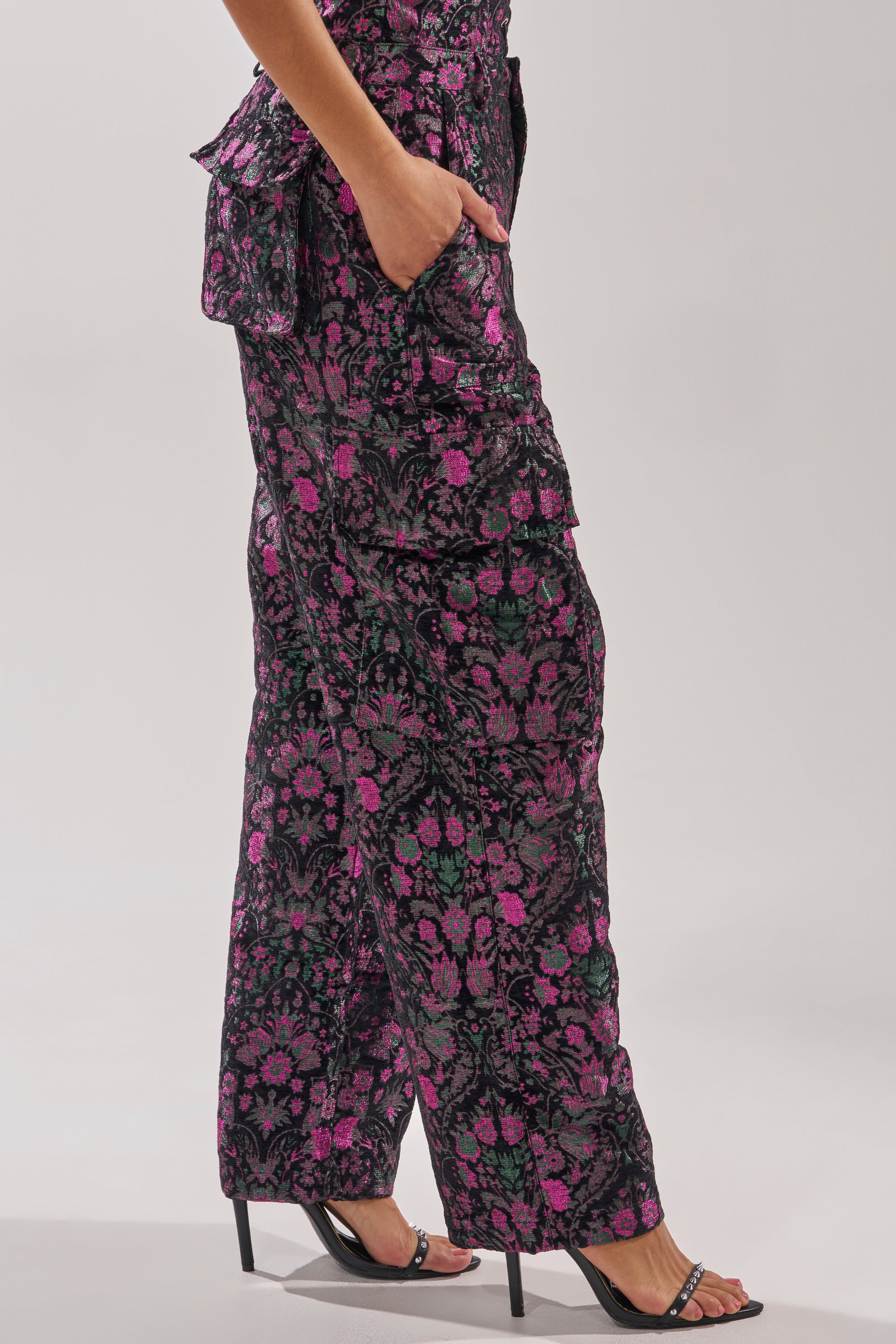 Wearing the KIMMY FLORAL TROUSER, a high-rise straight-leg pant with metallic floral details, and black heels, a person stands with one hand in their pocket against a plain background. 4