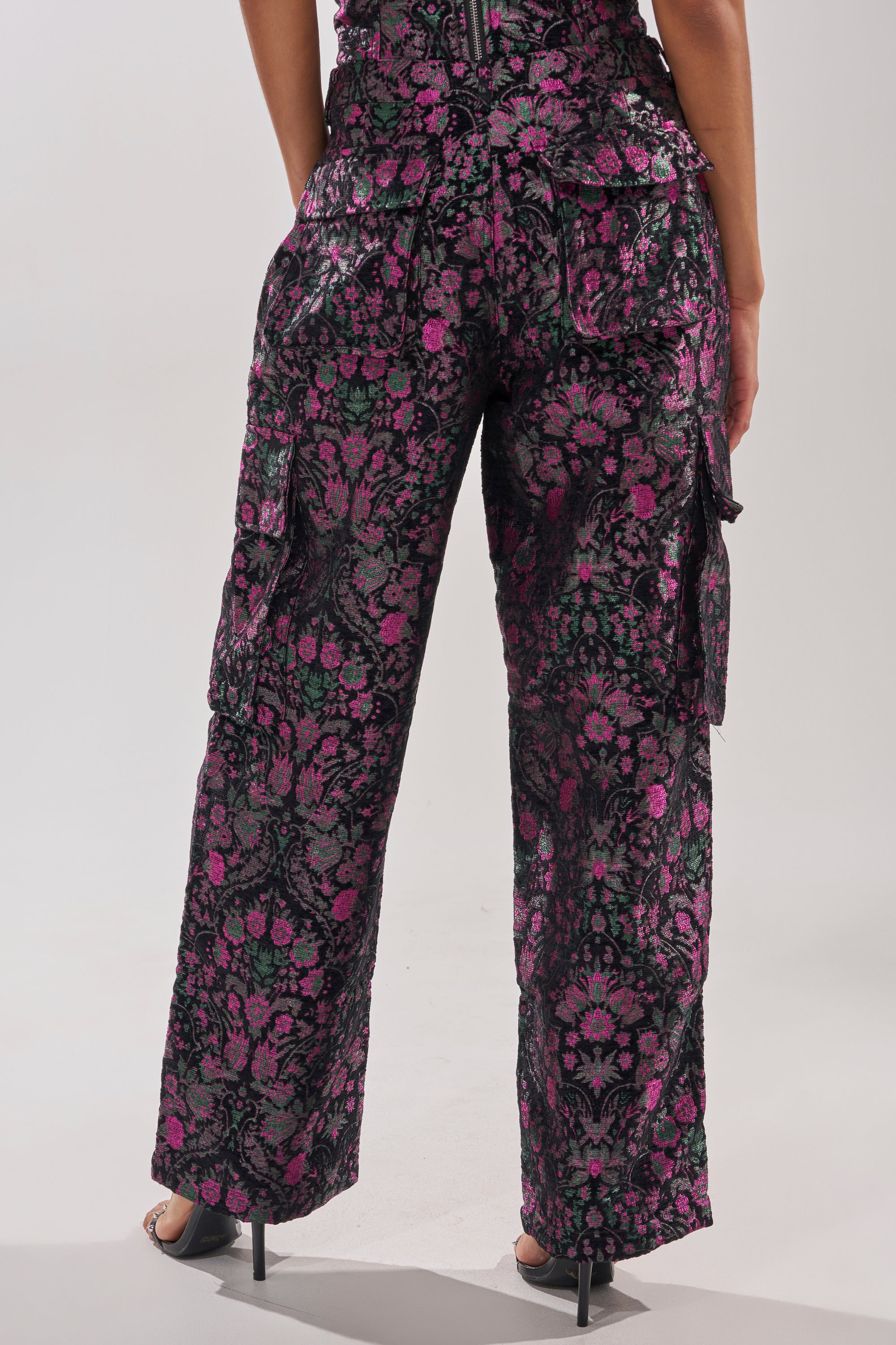 A person stands facing away, wearing the KIMMY FLORAL TROUSER featuring a high-rise fit and metallic floral details, paired with open-toed black heels. 5