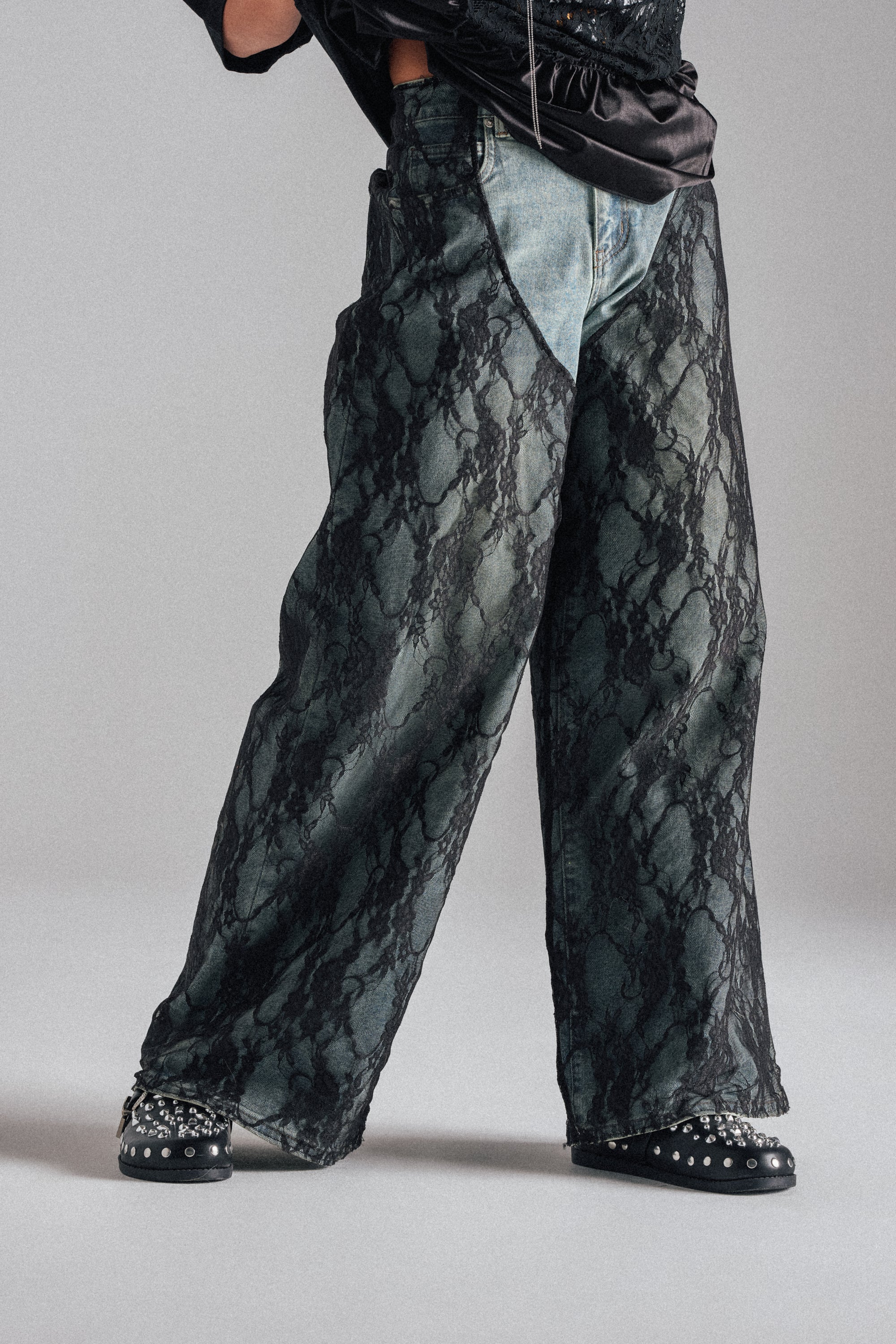 Wearing SEXY LACE CHAP DENIM high-rise, wide-leg jeans with a snake print overlay and pointed studded black shoes, the person stands against a plain gray backdrop. The upper body is mostly out of frame. 1