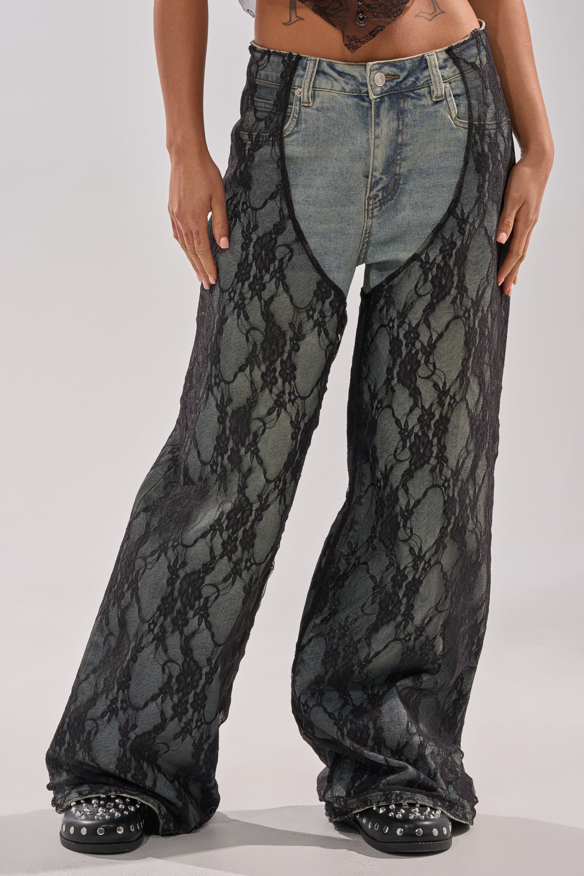 A person wears SEXY LACE CHAP DENIM—unique jeans with blue high-rise denim shorts in front and a black lace overlay. Black studded shoes and part of a tattooed torso complete the bold look. 0