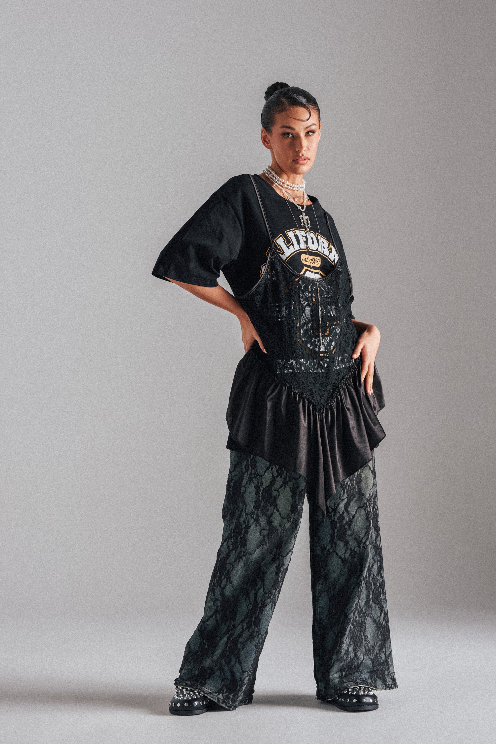 A woman poses confidently in SEXY LACE CHAP DENIM layered with a black oversize "California" tee, ruffled skirt, necklaces, earrings, and studded shoes for a bold and stylish look. 2