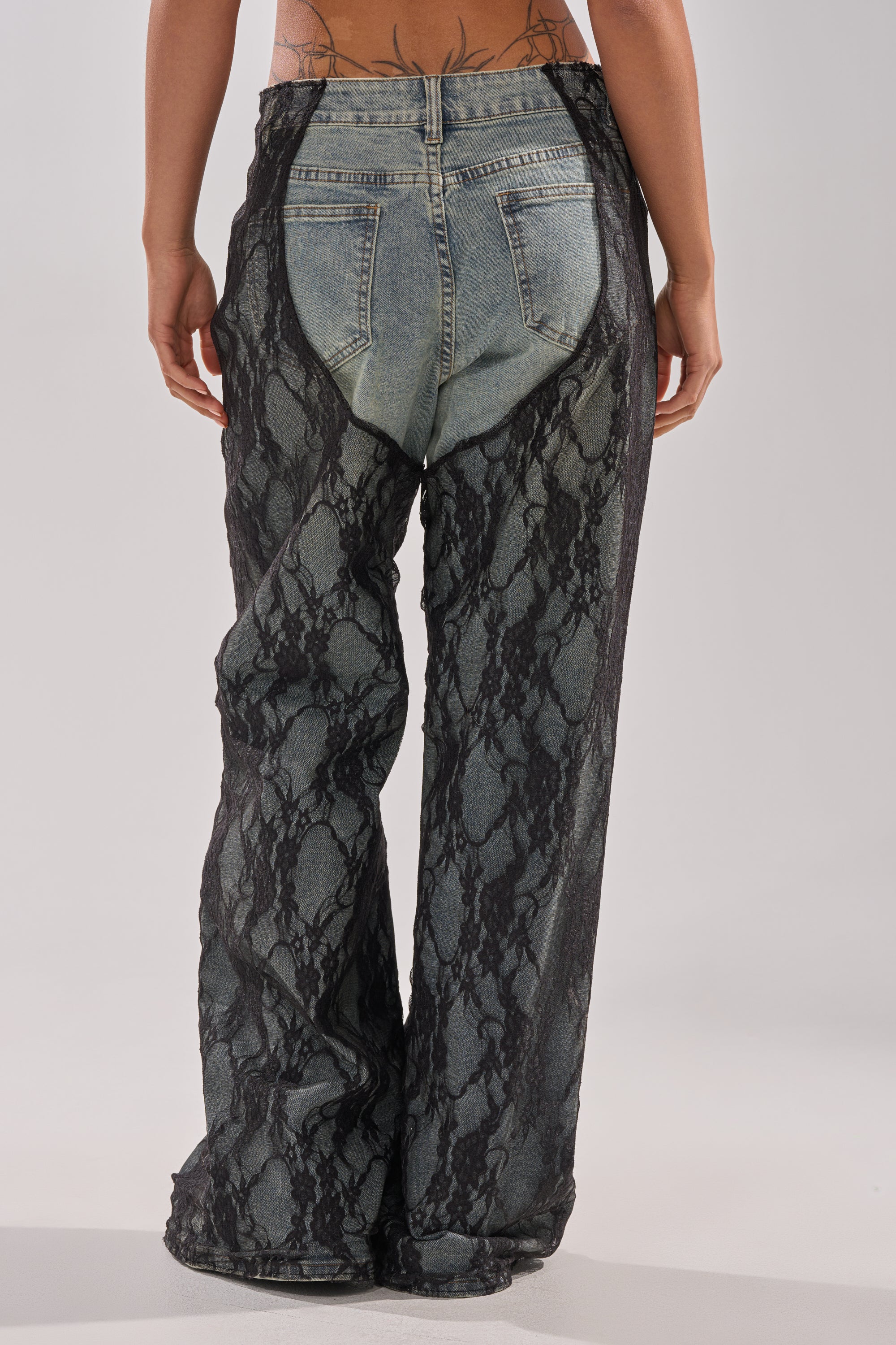 Person stands with their back to the camera, wearing SEXY LACE CHAP DENIM: light blue high-rise wide-leg jeans layered under sheer black floral lace chaps, revealing the jean style through the detailed lace. 5