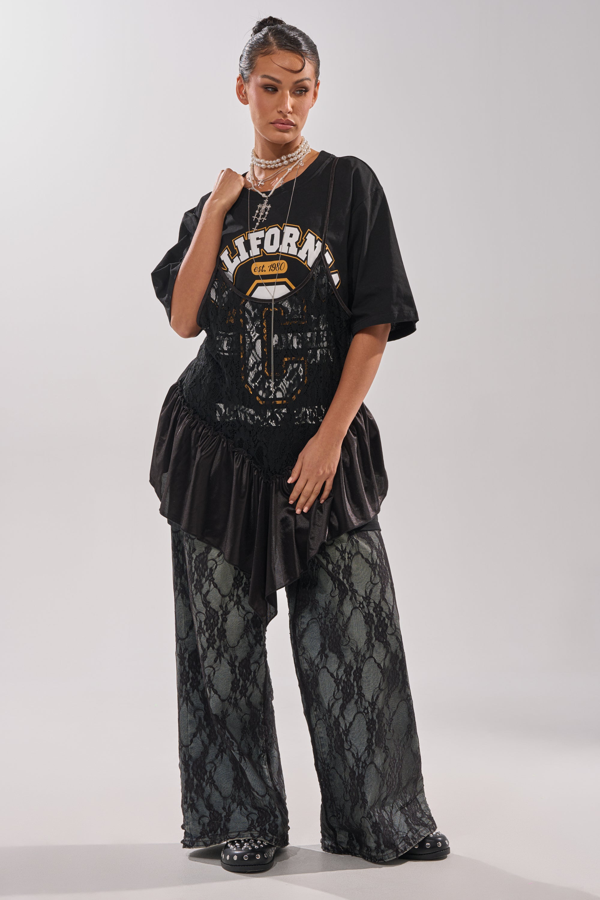 A woman poses in SEXY LACE CHAP DENIM, pairing the wide-leg jean floral lace pants with a black "California" tee, studded shoes, layered necklaces, a black lace corset, and a sleek updo. 7