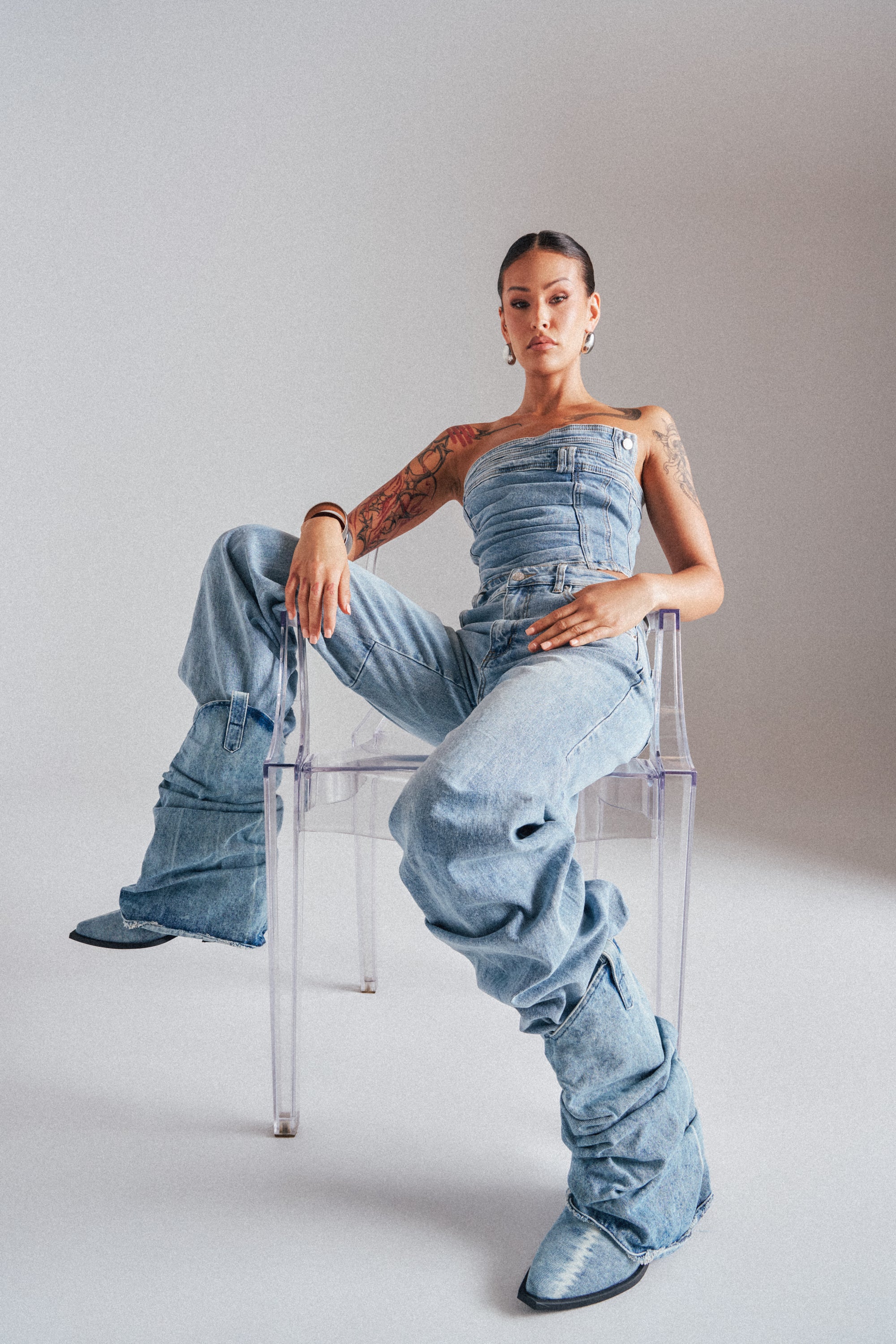 A tattooed individual sits confidently on a clear chair, wearing the ON YOUR MIND STACKED DENIM with boots. The plain, light-colored background enhances the style of this standout denim outfit. 0
