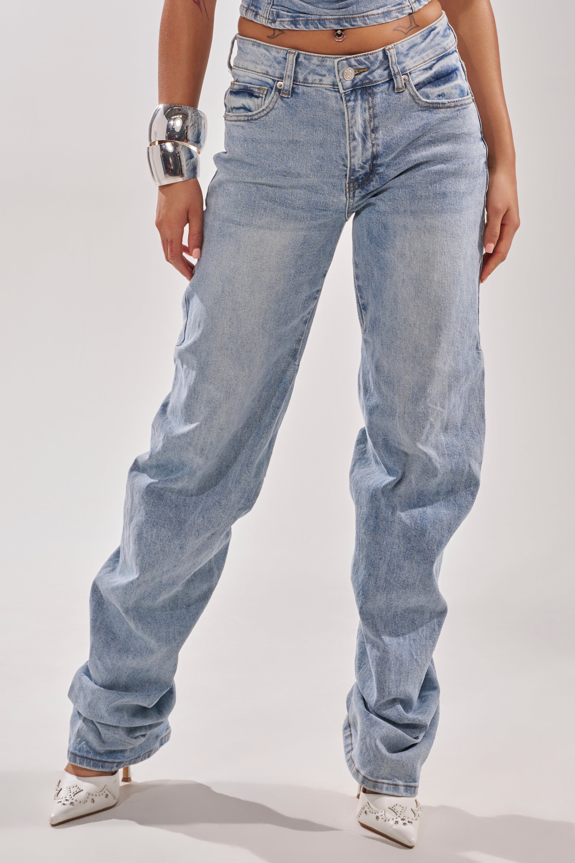 A person wearing ON YOUR MIND STACKED DENIM light blue high-rise baggy jeans with a matching crop top, white pointed-toe cut-out heels, and a chunky silver bracelet stands against a plain light background. 3