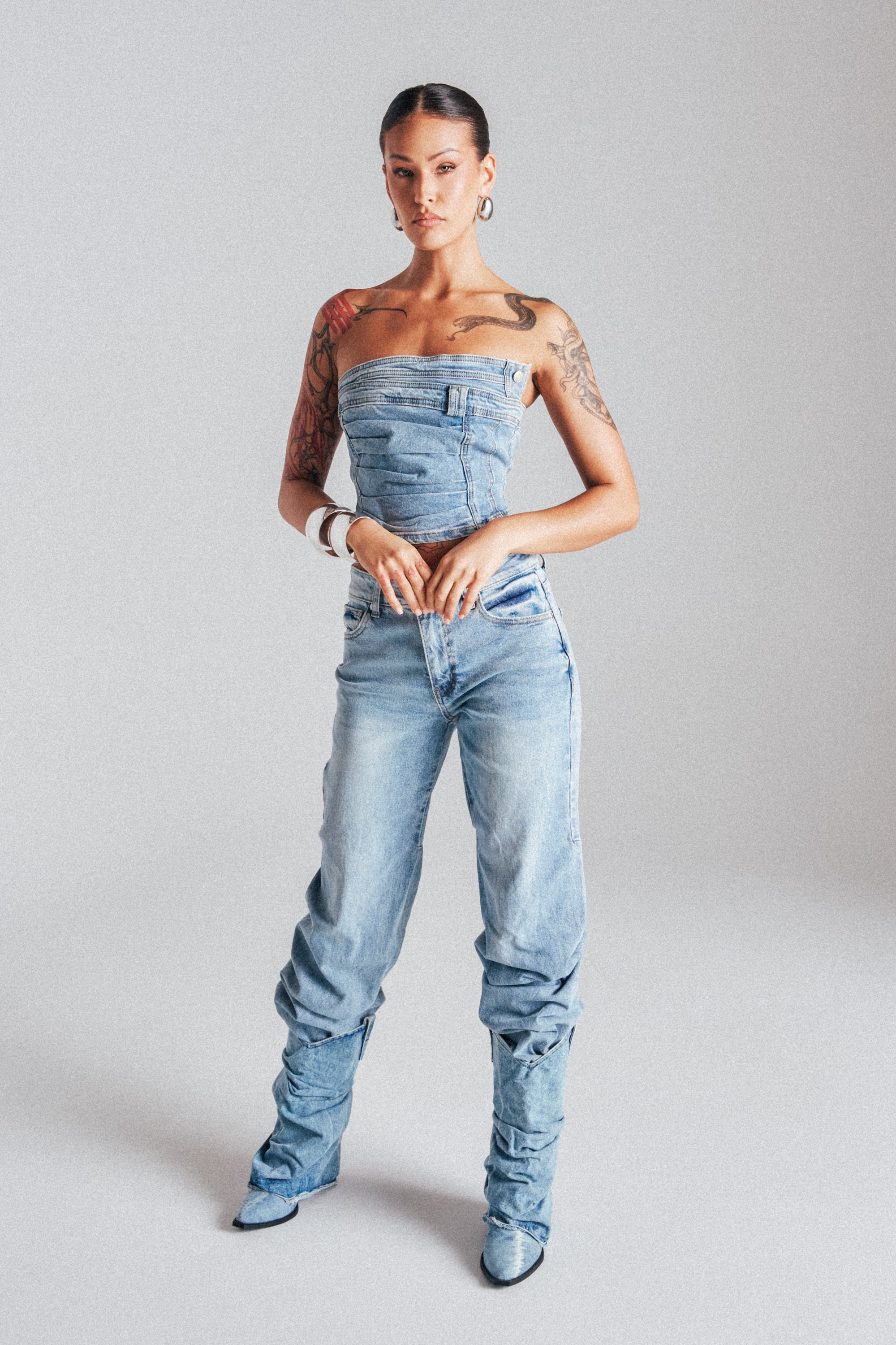 A tattooed woman poses confidently in the ON YOUR MIND STACKED DENIM strapless top, paired with matching high-waisted skinny jeans and pointed denim boots, against a plain light gray background. 1