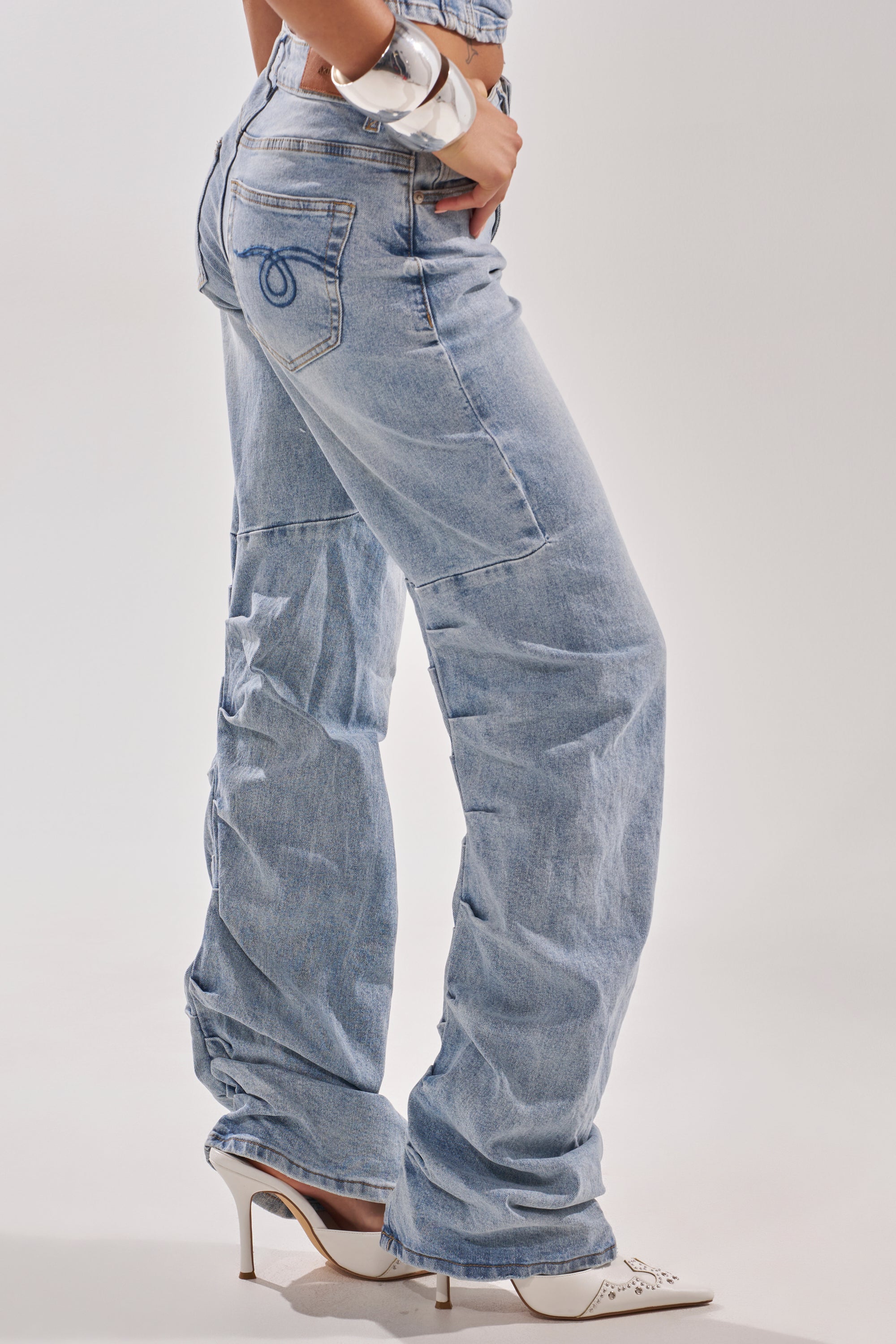 A person wears ON YOUR MIND STACKED DENIM, featuring light-wash, loose fit with back pocket details, paired with white high heels and a chunky white bracelet, standing against a plain light background. 5