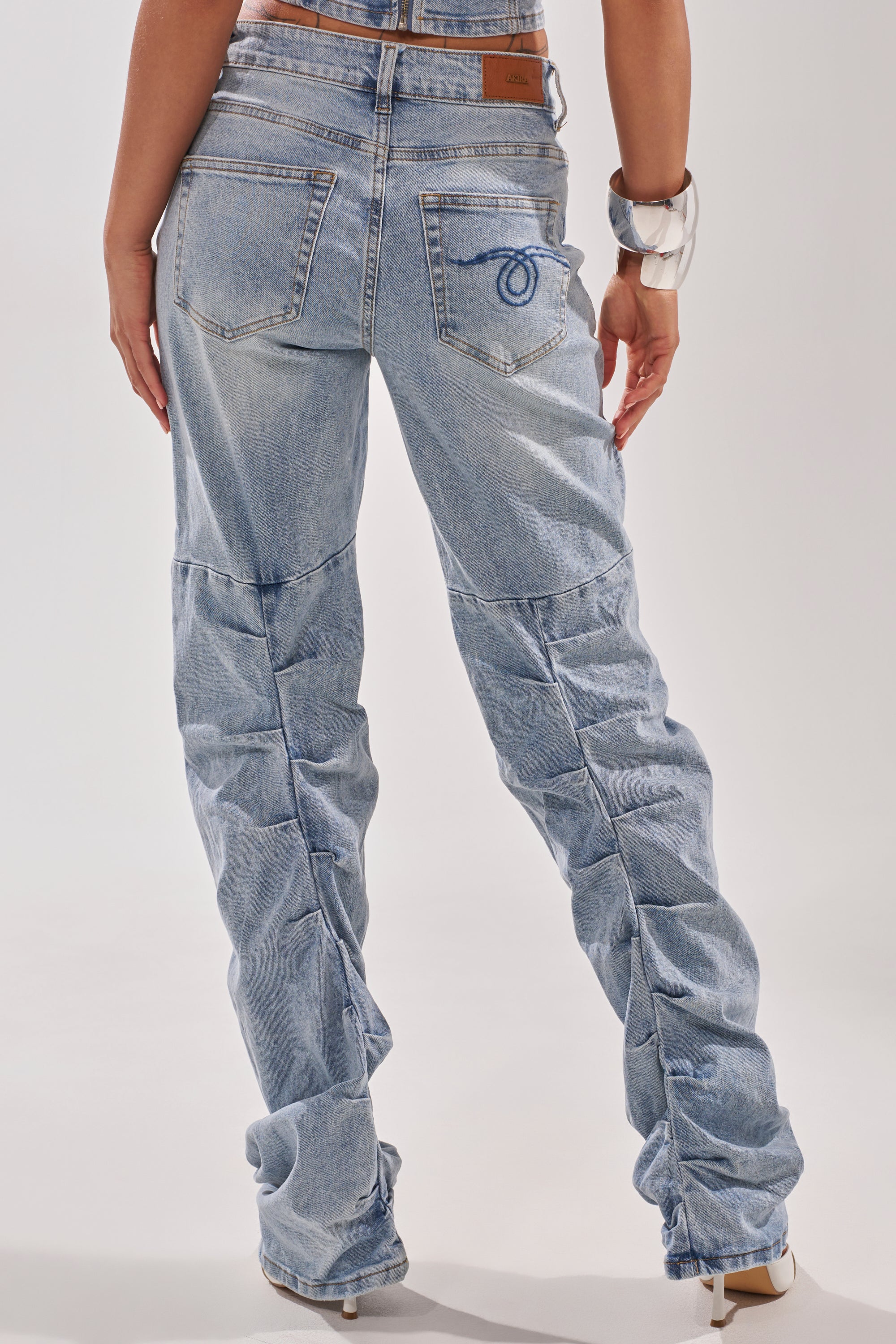 A person is shown from the back wearing ON YOUR MIND STACKED DENIM in light blue with a loose fit, paired with beige heeled shoes and a silver cuff bracelet, one hand resting near the back pocket. 6