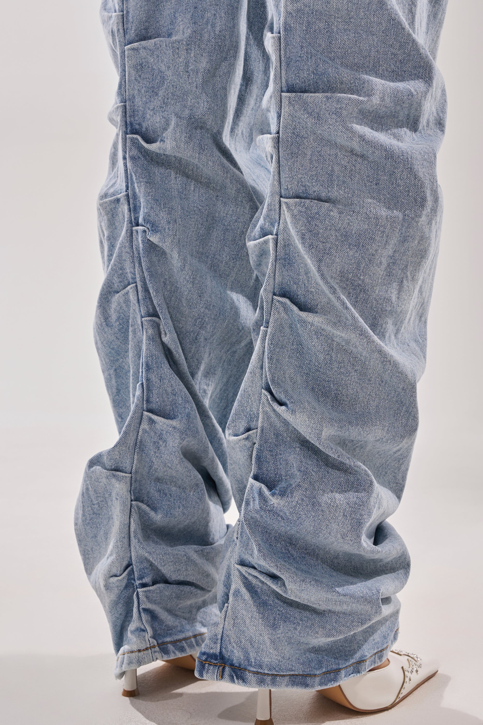 A close-up of a person wearing the ON YOUR MIND STACKED DENIM—light blue, high-rise ruched jeans with a stacked look—paired with white heeled shoes on a light-colored surface. 9