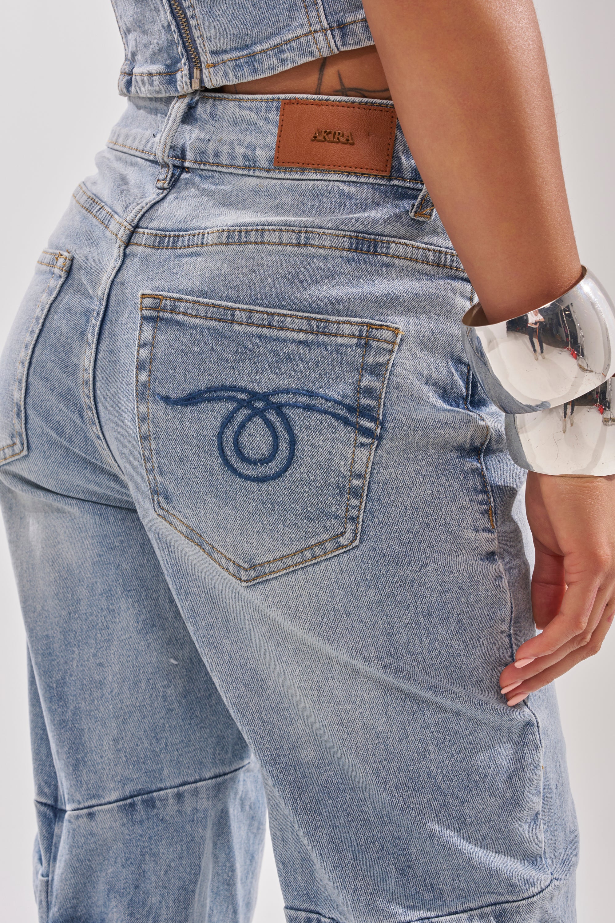 A person wearing ON YOUR MIND STACKED DENIM—light blue, high-rise jeans with dark blue stitched back pockets and a tan leather patch—accessorized with chunky silver bangles. 10