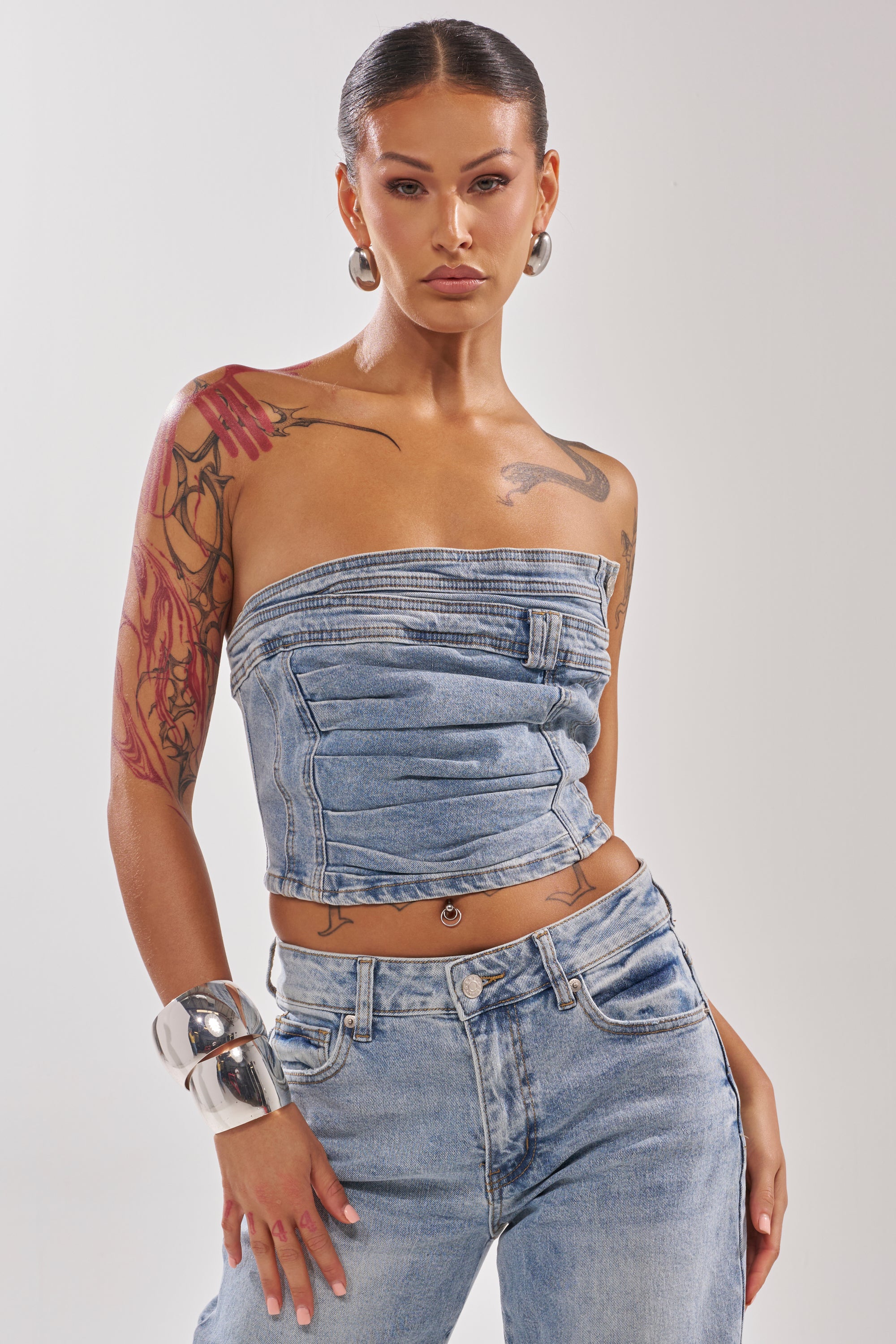 A woman with slicked-back hair and bold makeup models the ON YOUR MIND DENIM CORSET—a strapless, cropped, form-fitting denim top—paired with matching jeans, accessorized by large hoops and chunky silver bracelets against a plain background.