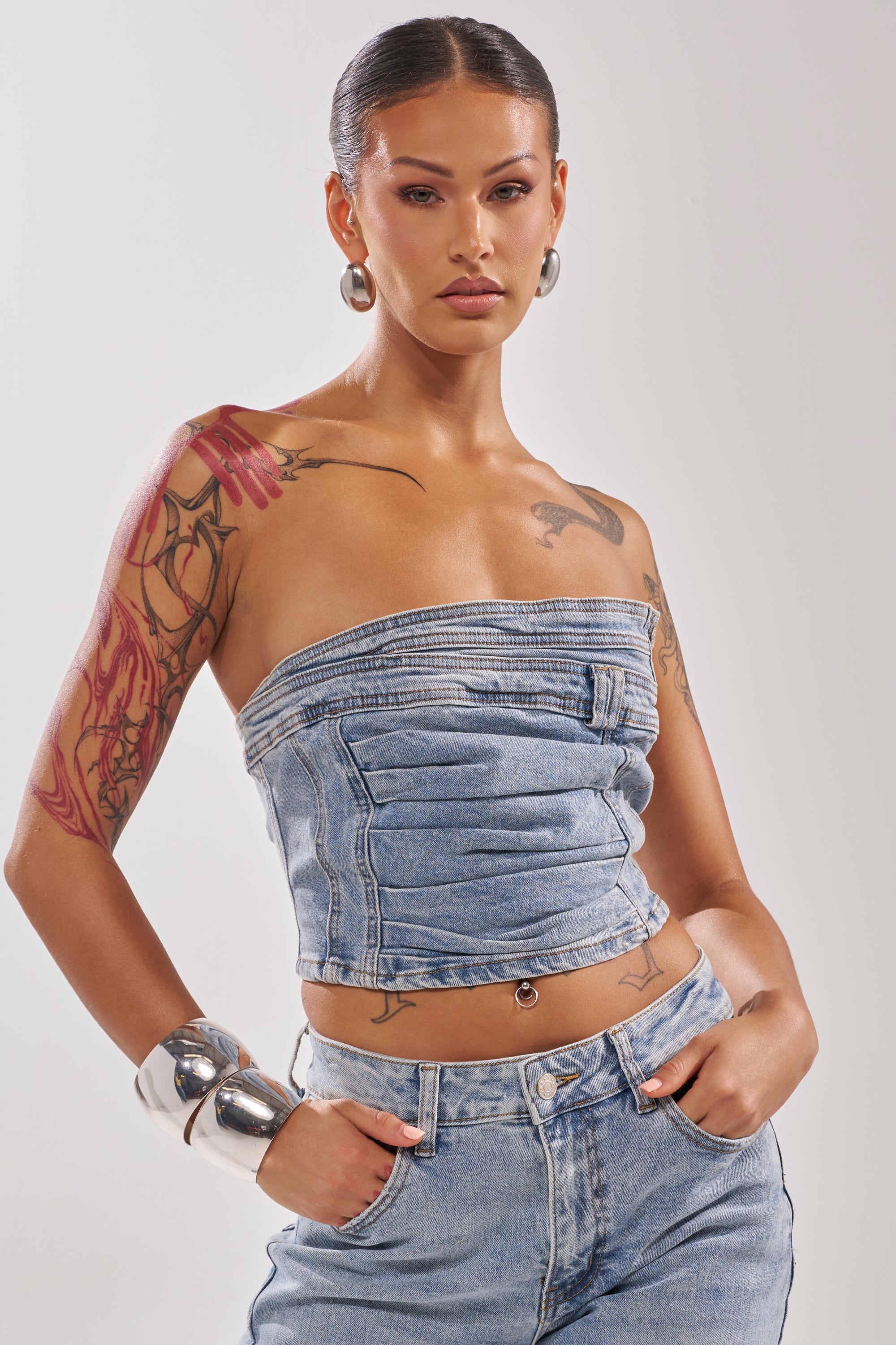A person with slicked-back hair wears the ON YOUR MIND DENIM CORSET and high-waisted jeans. Sporting tattoos, large hoop earrings, and silver bangles, they pose with hands in pockets against a plain background.