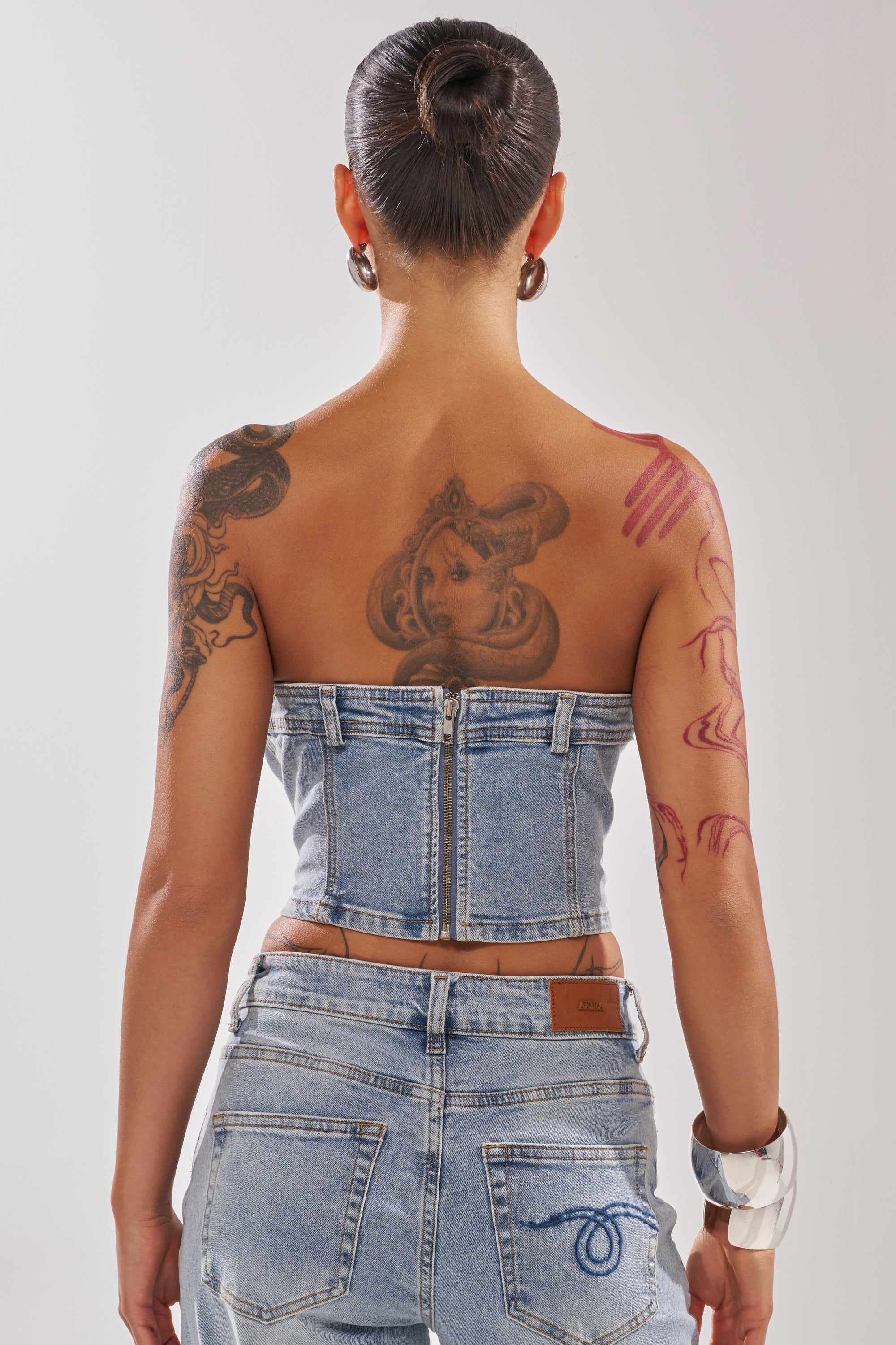A person with their hair in a bun and tattoos on their upper back and arm wears the ON YOUR MIND DENIM CORSET and jeans, accessorized with a chunky silver bracelet, facing away from the camera.