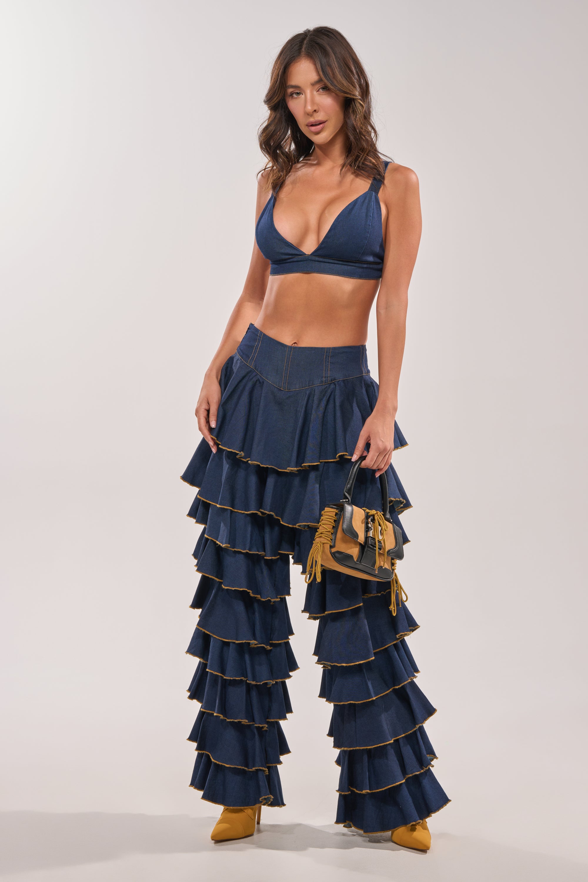 A woman poses confidently in SHAKE IT OUT DENIM RUFFLE JEANS and a deep blue bralette, accessorized with a brown fringe handbag and yellow heels, set against a plain light background. 0