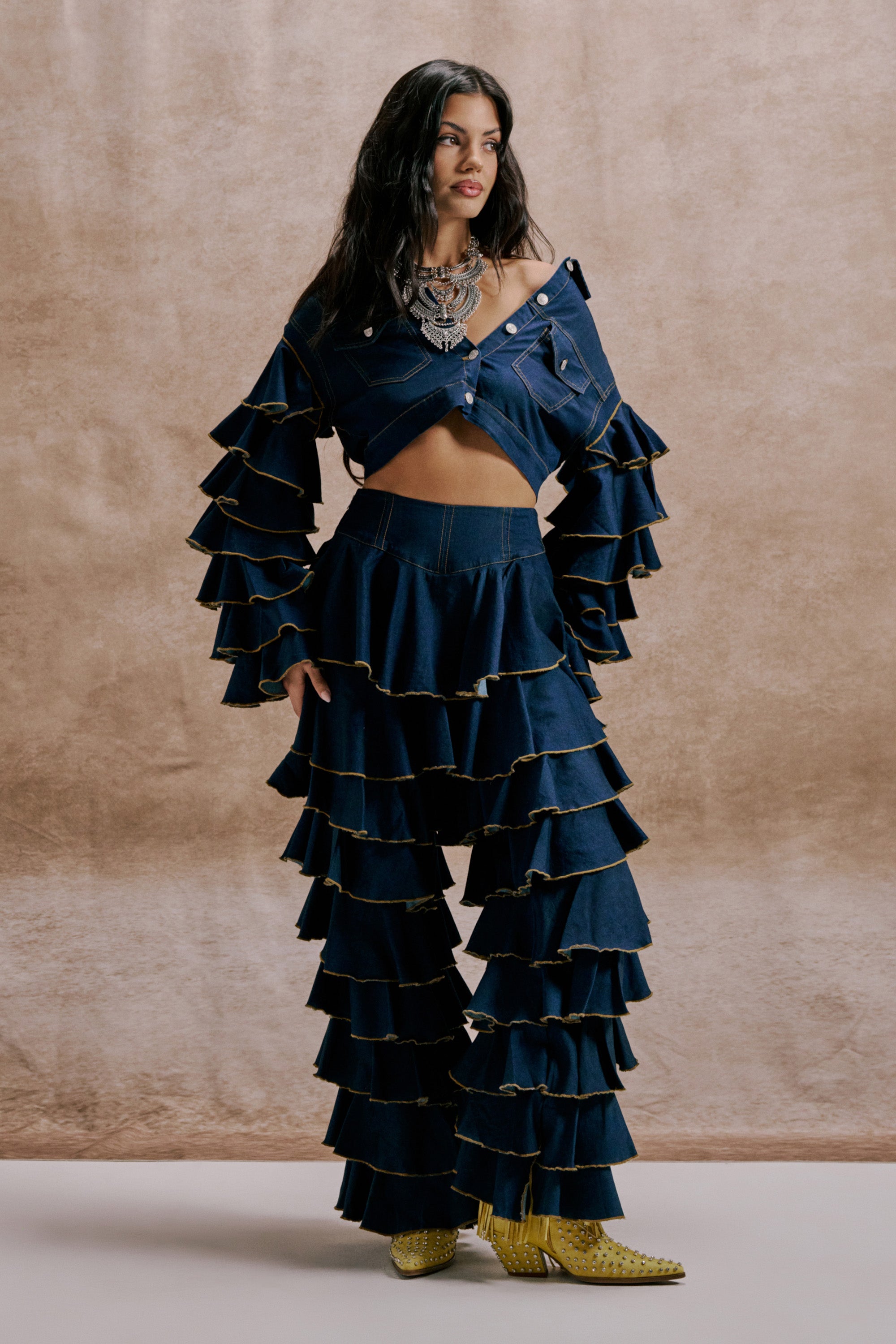 A woman poses in SHAKE IT OUT DENIM RUFFLE JEANS featuring high-waisted denim with blue ruffled layered sleeves, styled with bold jewelry and yellow studded boots, against a neutral backdrop. 1
