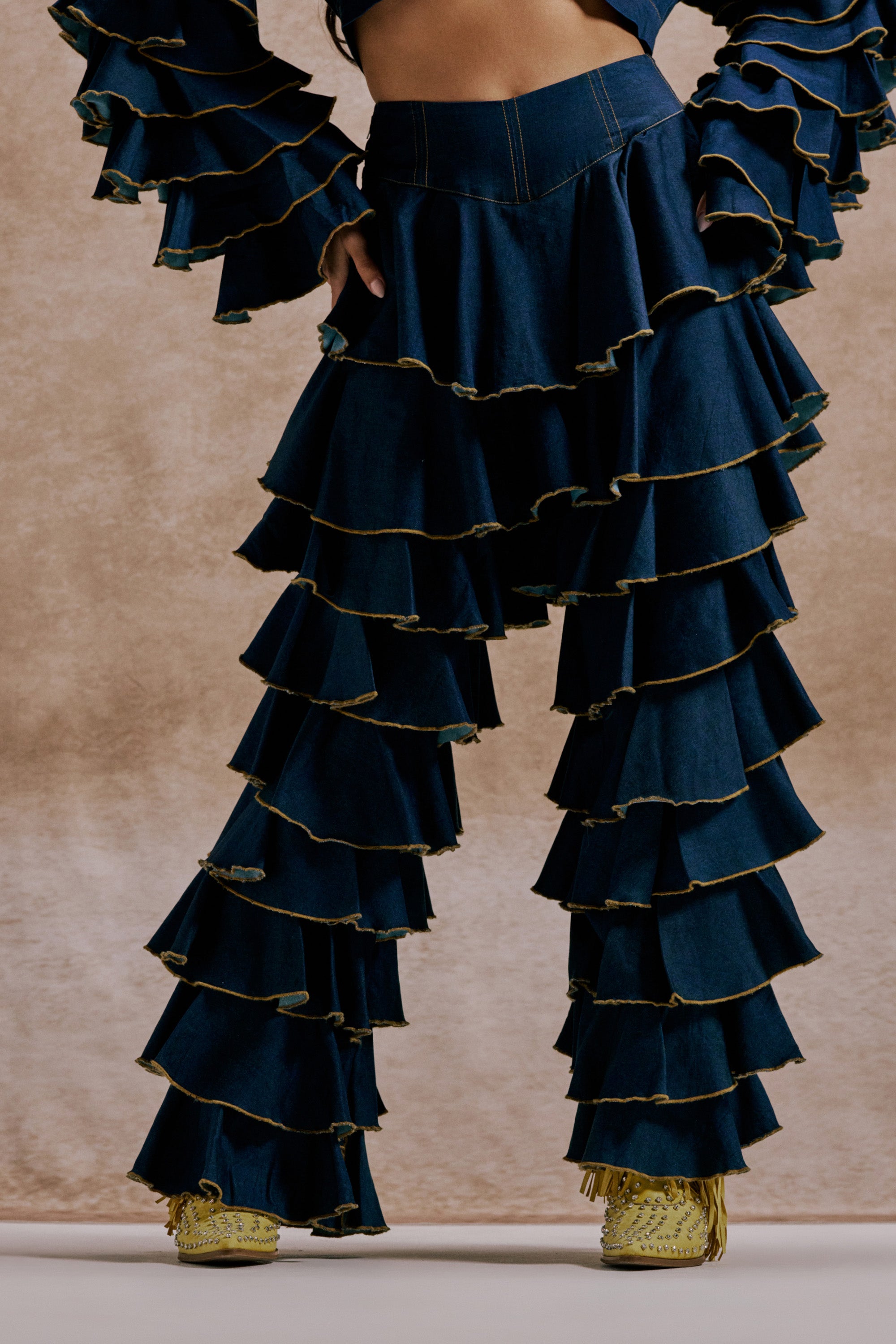 A person stands against a neutral background wearing SHAKE IT OUT DENIM RUFFLE JEANS, featuring high-waisted design and multiple ruffled layers, paired with yellow shoes. 2