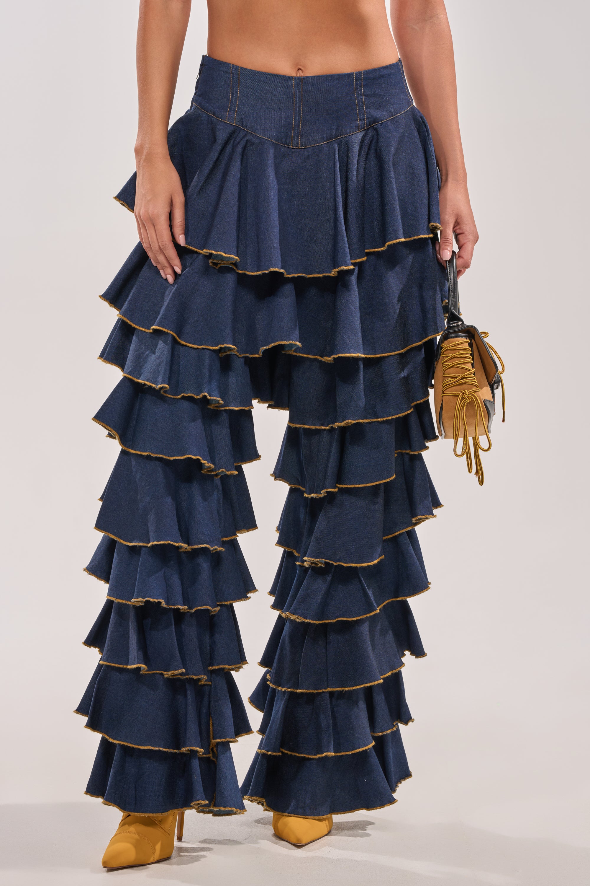 Wearing SHAKE IT OUT DENIM RUFFLE JEANS in dark blue with tiered ruffles, the person holds a small mustard yellow handbag and wears matching pointed-toe shoes. The photo is cropped at the torso. 4