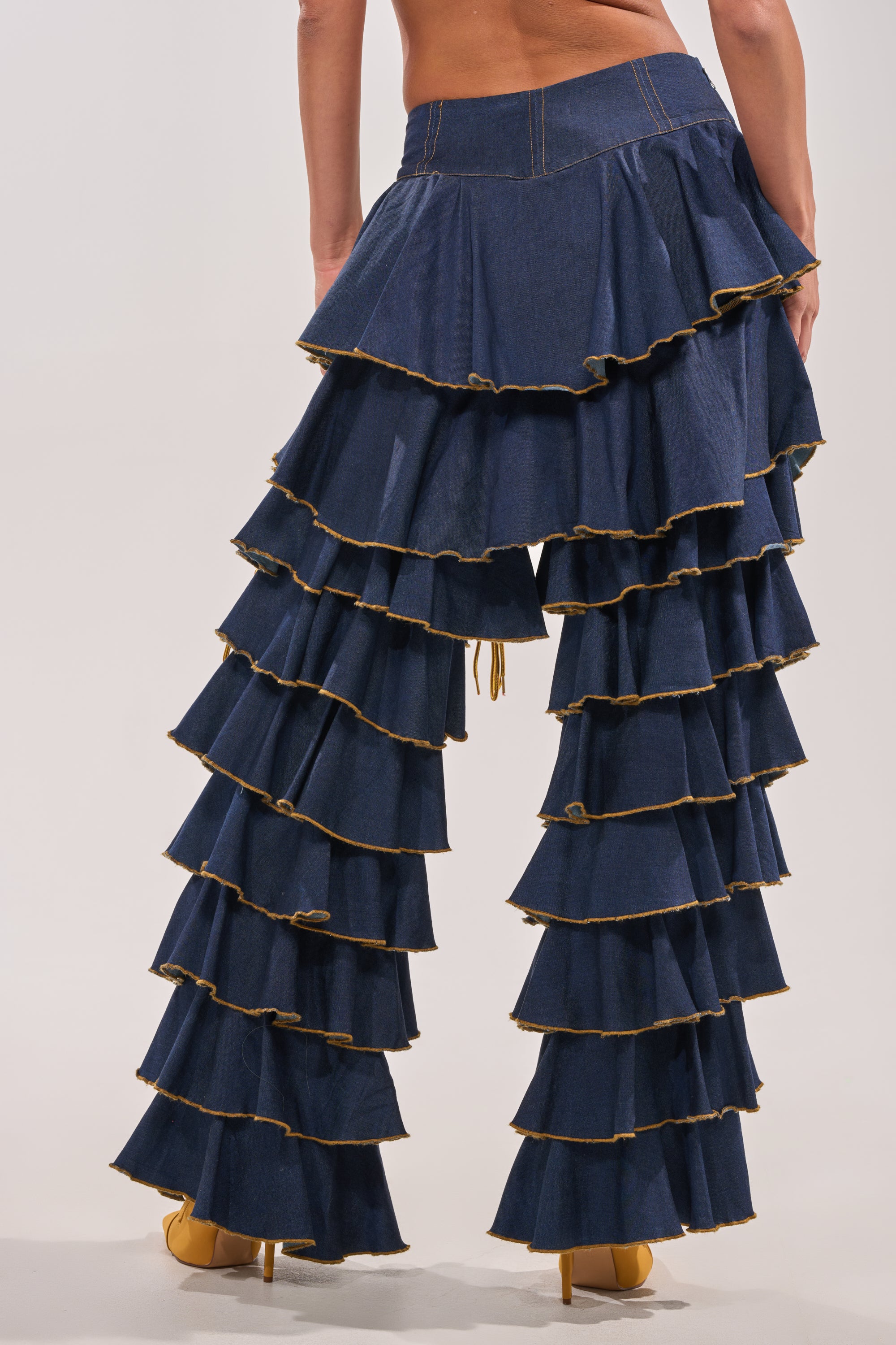 A person models the SHAKE IT OUT DENIM RUFFLE JEANS, featuring dramatic ruffle tiers and yellow stitching, paired with yellow high-heeled shoes. The bold, high-waisted denim look is photographed from behind against a plain light background. 6