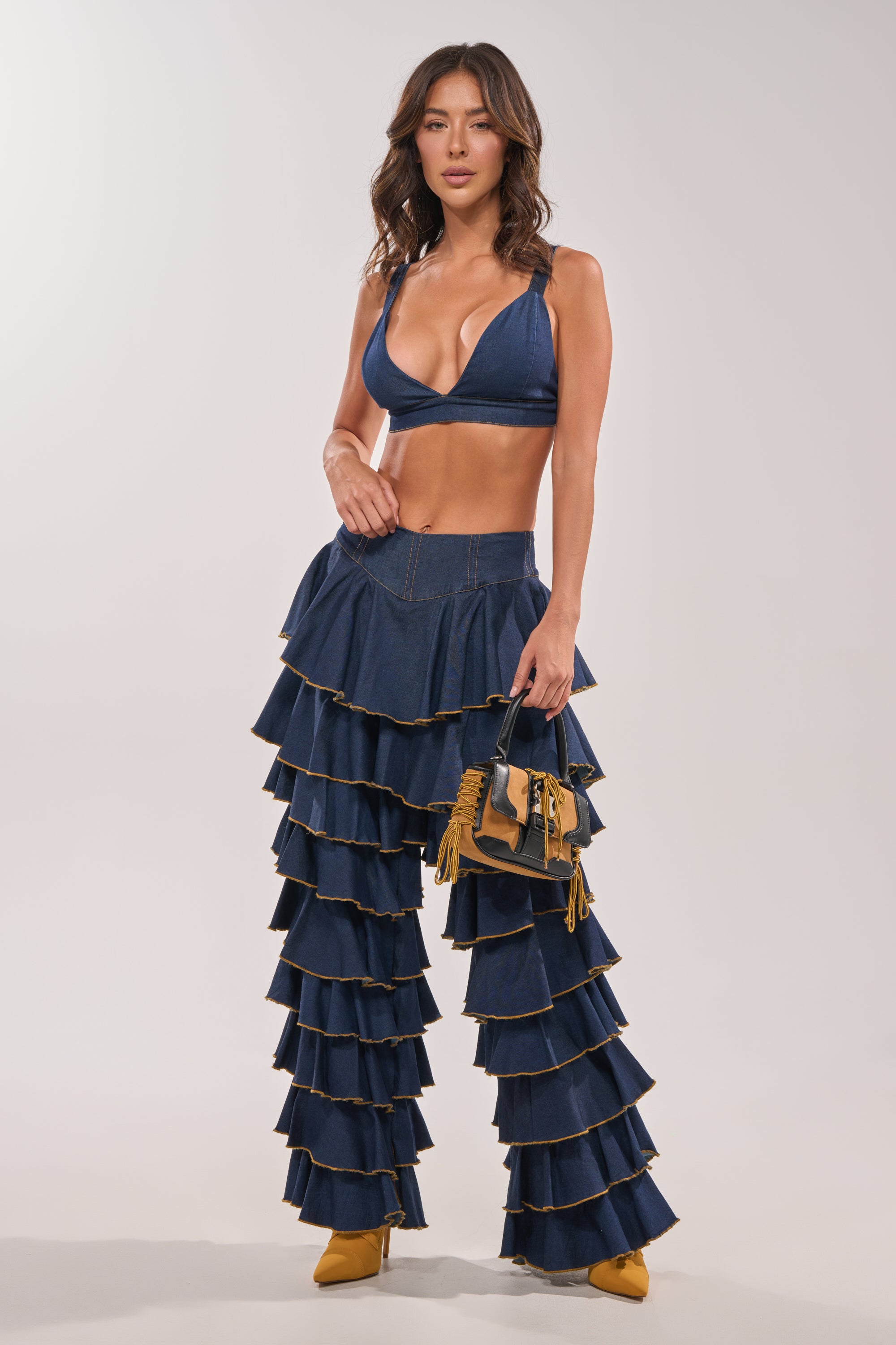A woman wears SHAKE IT OUT DENIM RUFFLE JEANS with a deep blue denim bralette, holding a small fringed handbag and standing in yellow pointed heels against a plain light background. 7