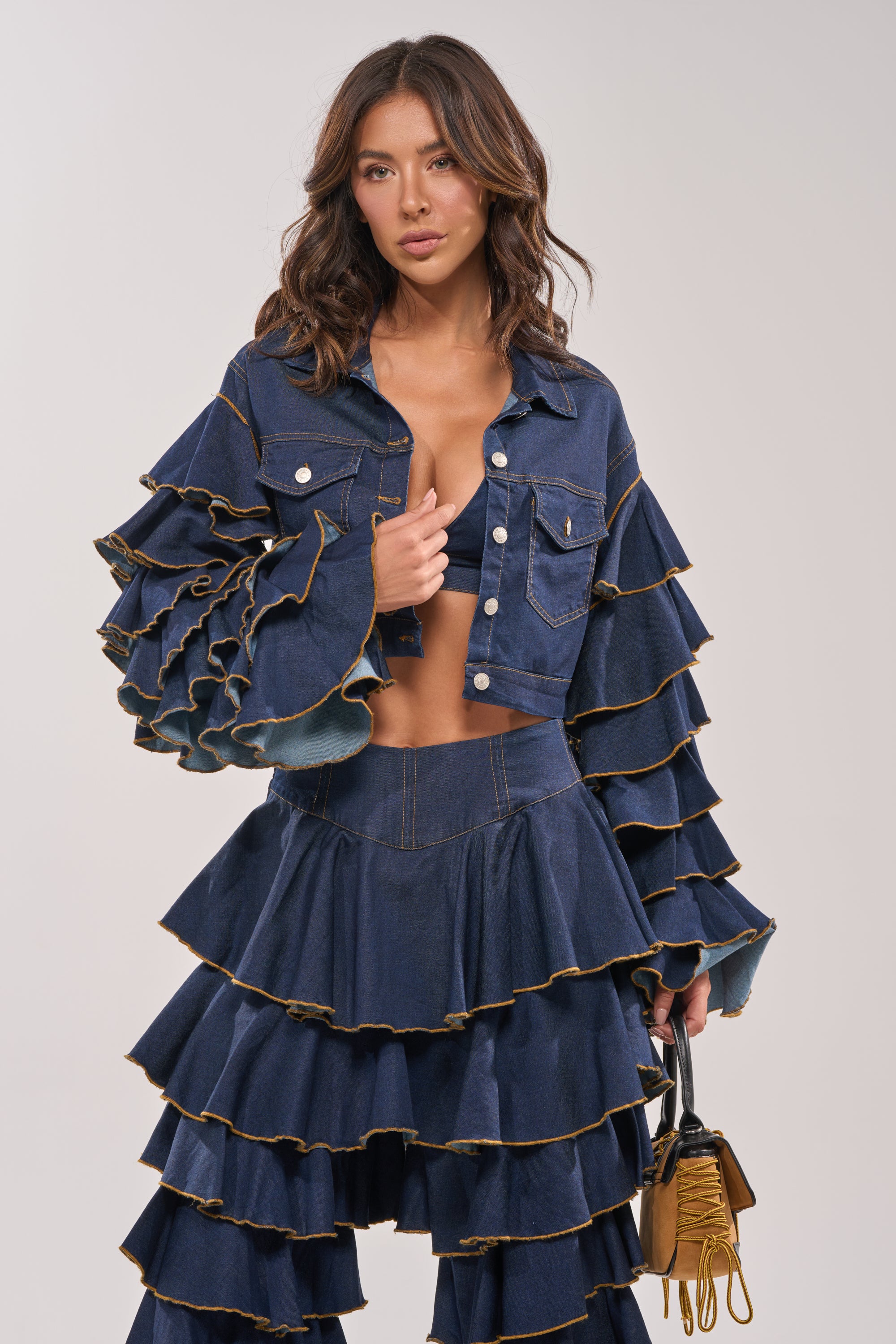 Wearing the SHAKE IT OUT CROPPED DENIM JACKET with tiered sleeves and a ruffled skirt, she stands against a plain background, holding a small yellow handbag—creating an effortless statement layered look. 3