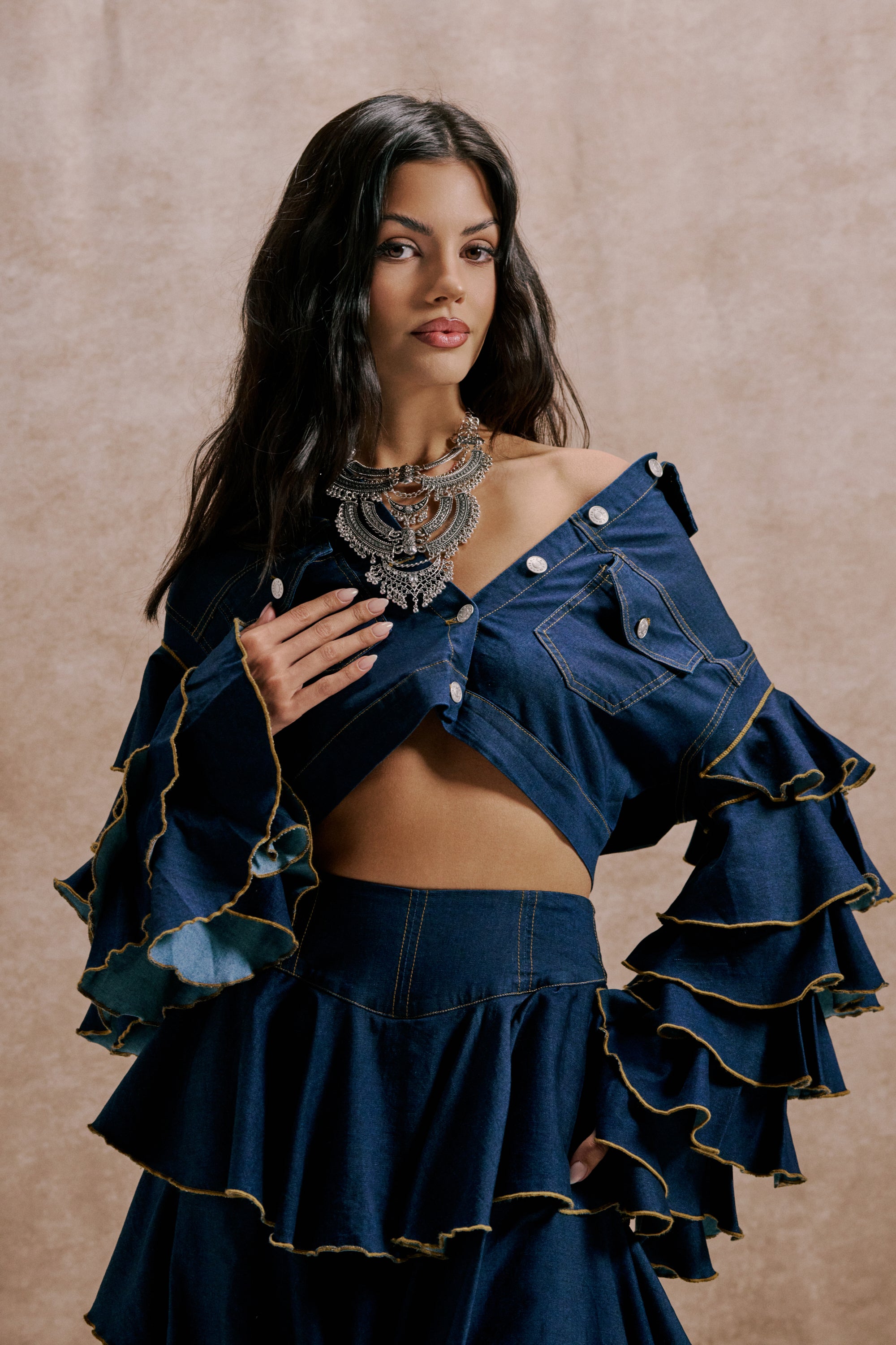 A woman poses in the SHAKE IT OUT CROPPED DENIM JACKET, featuring ruffled, off-shoulder design and yellow stitching, styled with an ornate silver necklace for a bold look against a neutral beige backdrop. 1