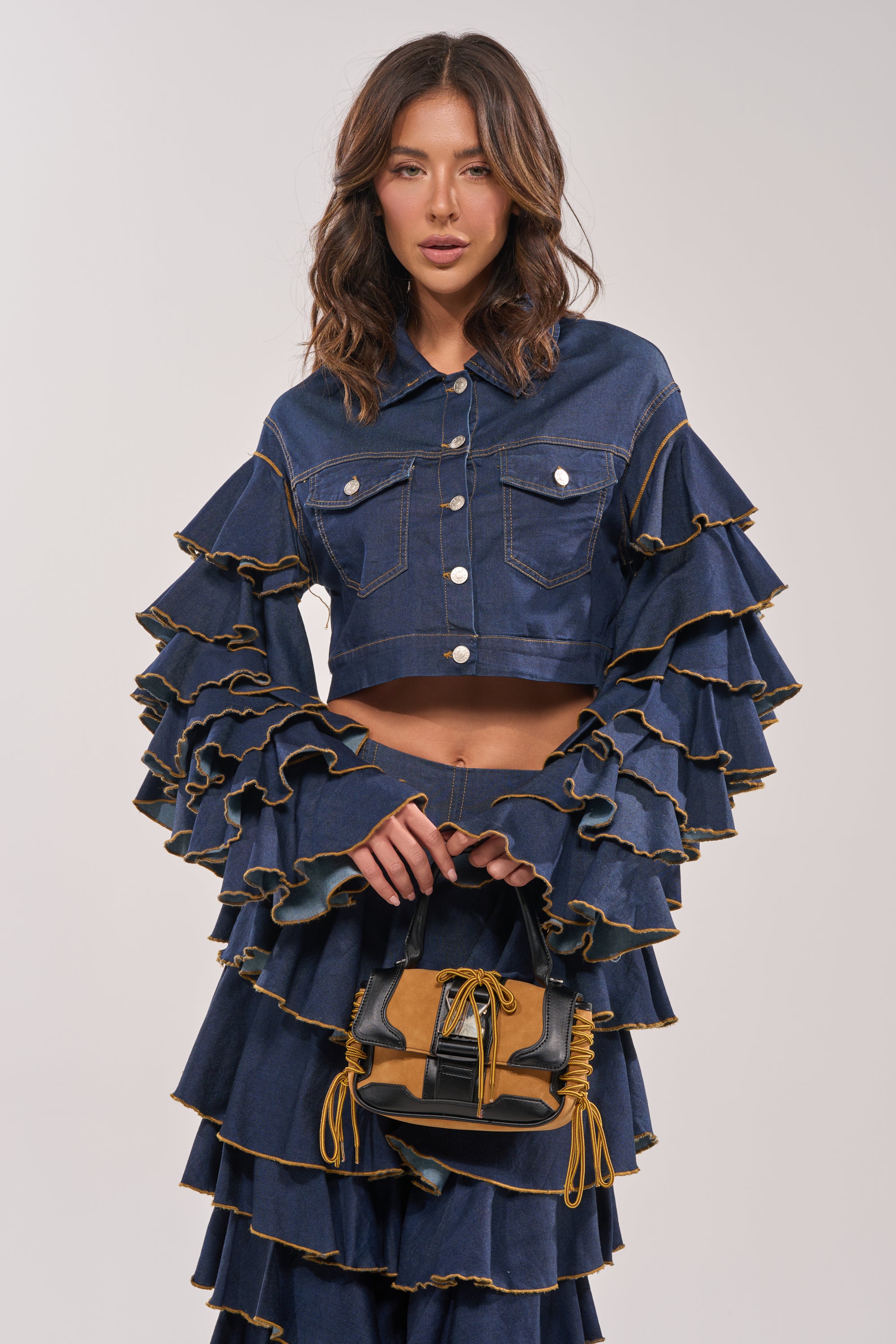A woman models the SHAKE IT OUT CROPPED DENIM JACKET with tiered sleeves and yellow stitching, paired with a matching skirt and a small brown-and-black handbag, against a plain light background for bold layered style. 4