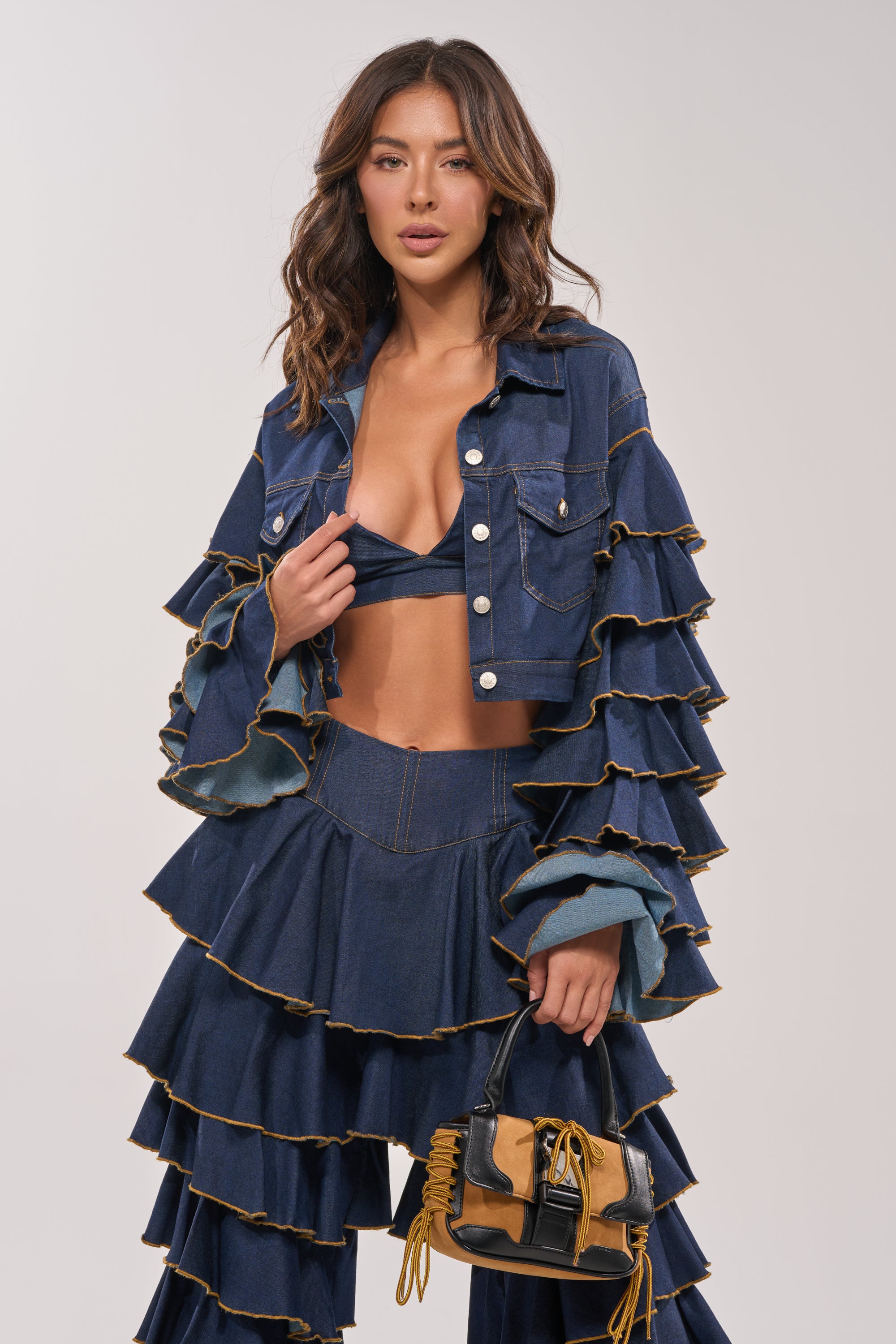 A woman poses confidently in the SHAKE IT OUT CROPPED DENIM JACKET with tiered sleeves and matching bralette, paired with denim pants and a tan and black handbag, set against a plain, light background. 5