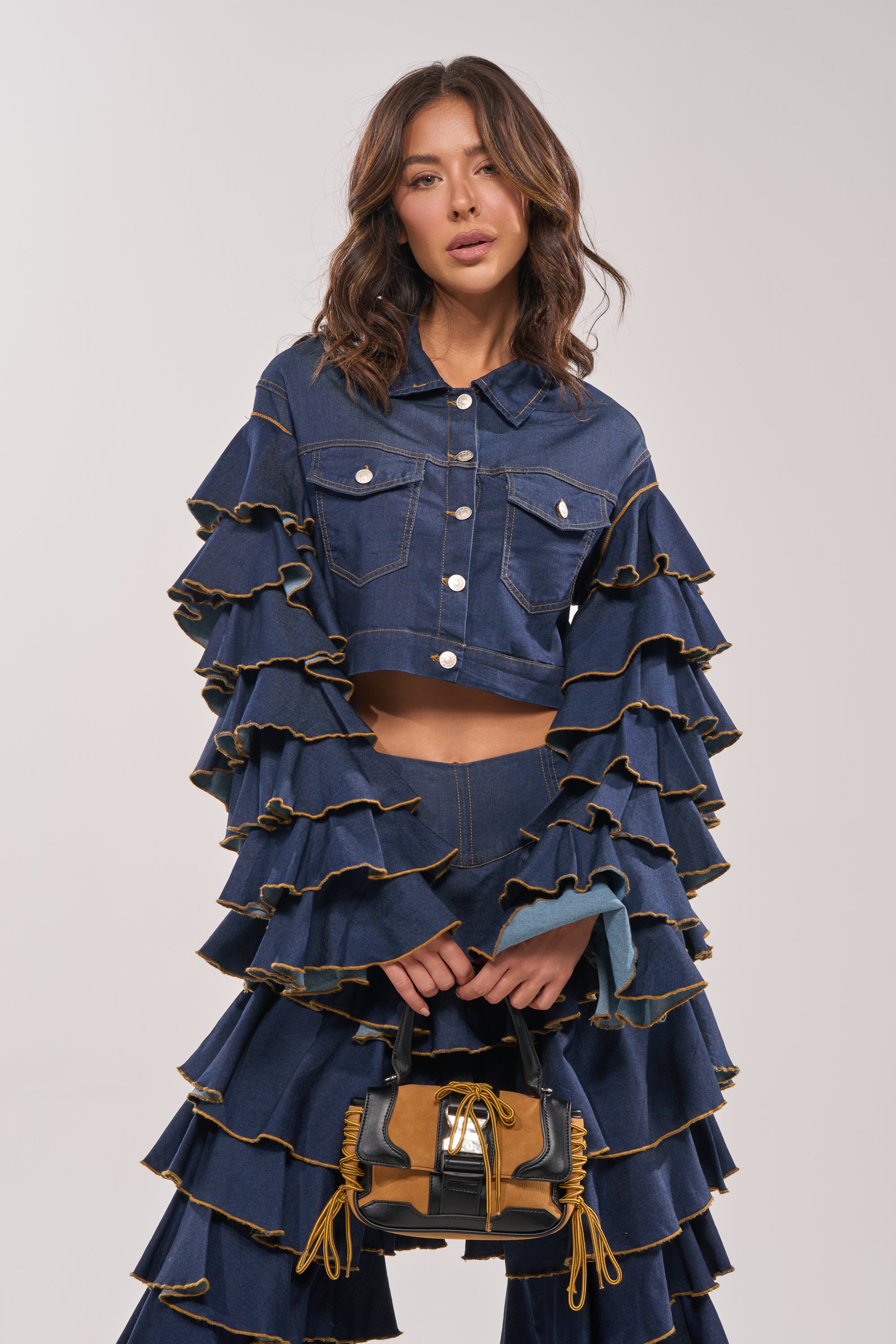A woman models the SHAKE IT OUT CROPPED DENIM JACKET with tiered sleeves and bold layering, pairing it with a small brown-and-black handbag with yellow accents against a plain light backdrop. 6