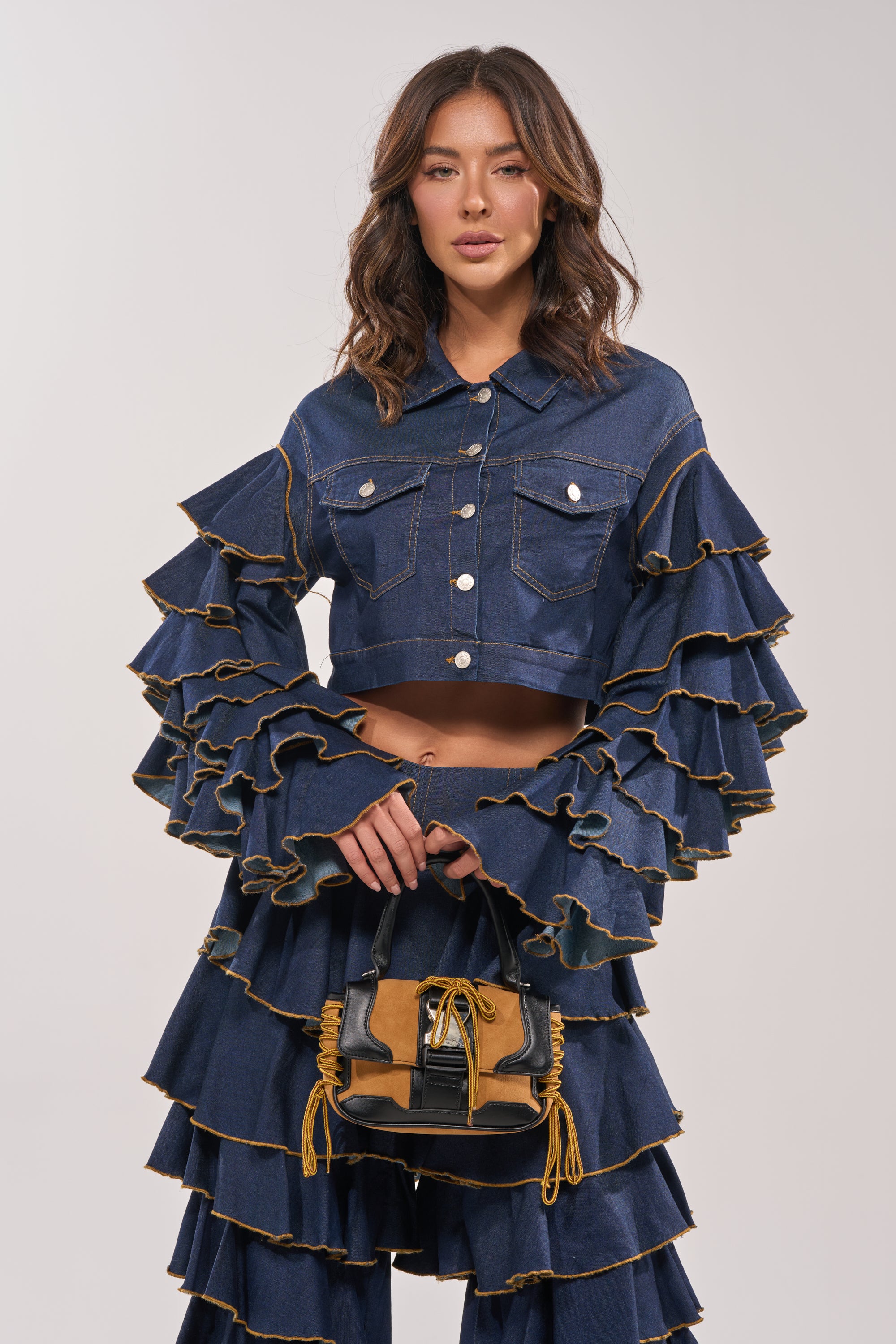 A woman wears the SHAKE IT OUT CROPPED DENIM JACKET, featuring tiered sleeves and layered dark denim pants, holding a small handbag with yellow accents against a plain light background. 7