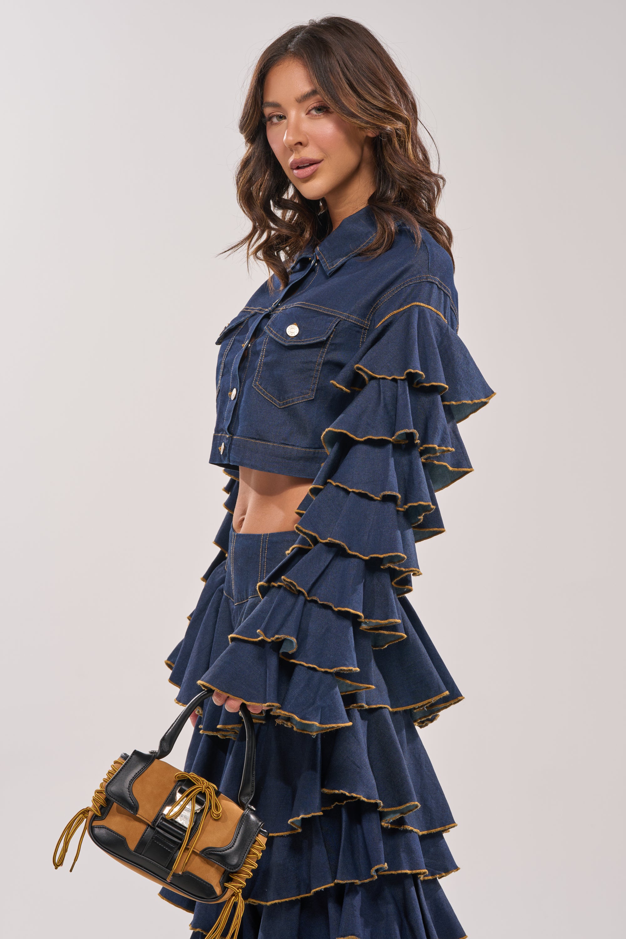 A woman models the SHAKE IT OUT CROPPED DENIM JACKET, a dark, ruffled piece with tiered sleeves, paired with a flared skirt and a brown-and-black tassel handbag—confidently posing for statement layering. 8