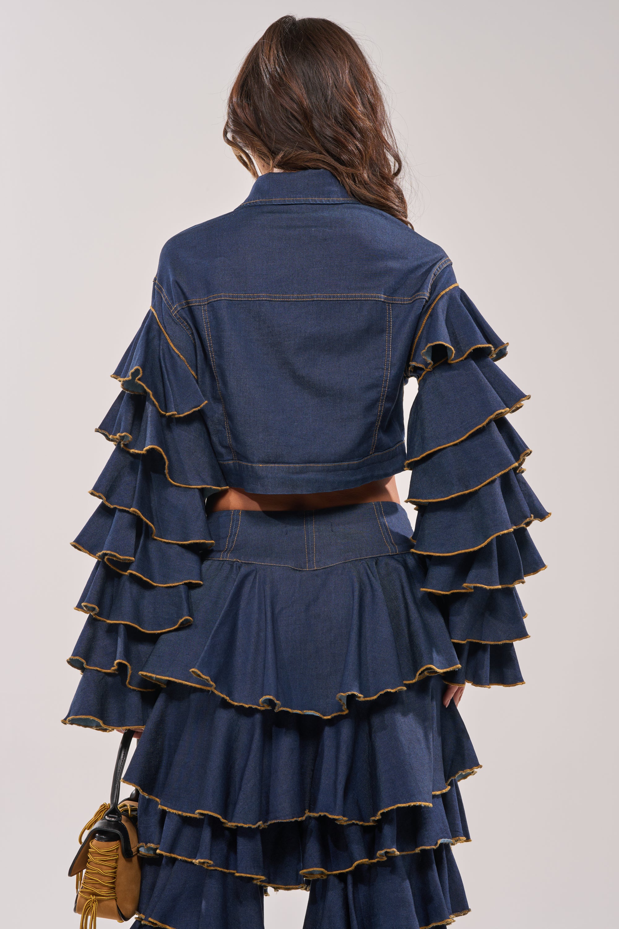 A woman with long, wavy brown hair stands facing away in the SHAKE IT OUT CROPPED DENIM JACKET with tiered sleeves and a ruffled skirt. She holds a small bag against a plain, neutral background. 9