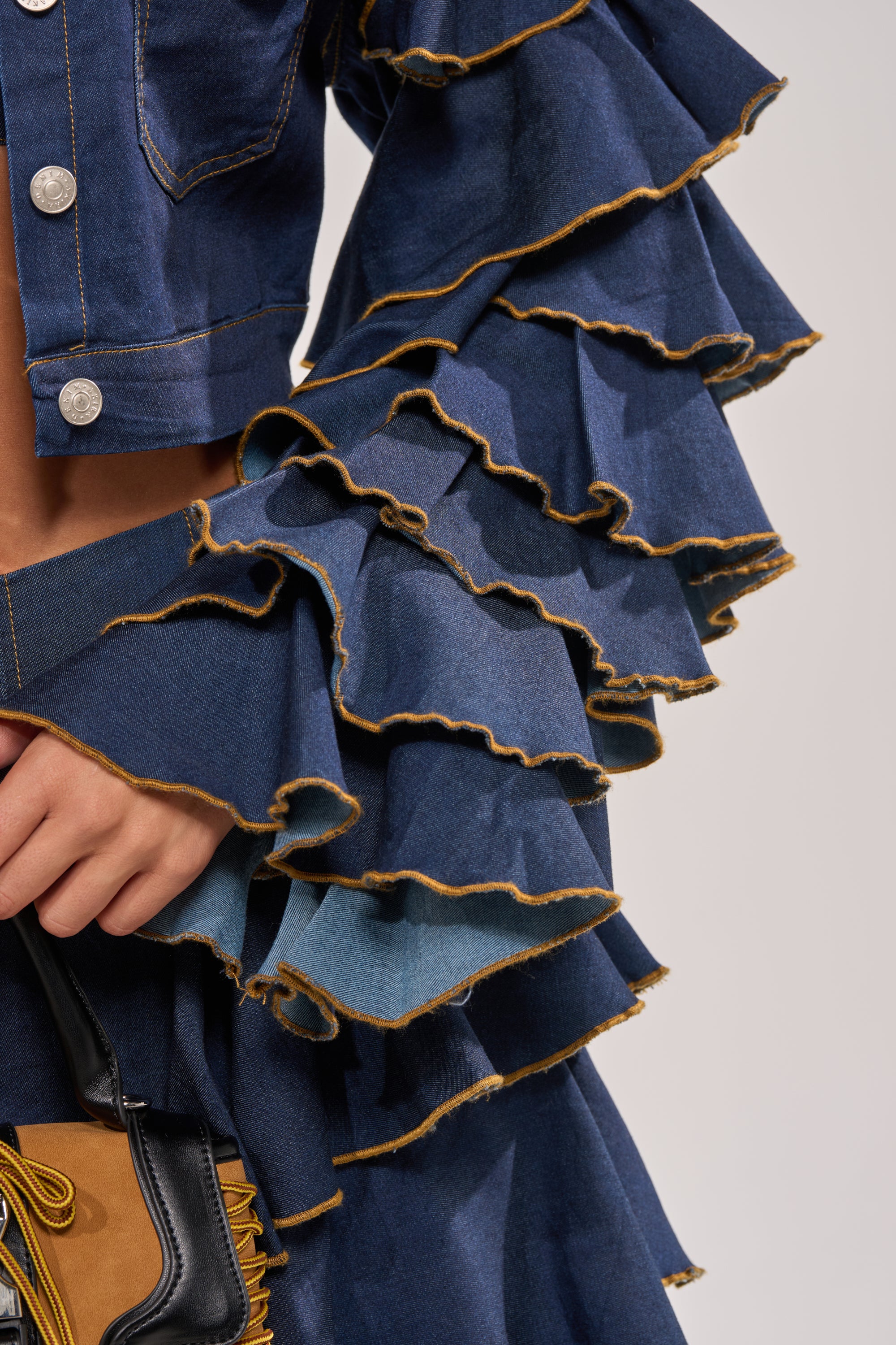 Close-up of a person wearing the SHAKE IT OUT CROPPED DENIM JACKET, featuring tiered sleeves and yellow stitching. They hold a small fringed bag with leather accents against a neutral background. 10