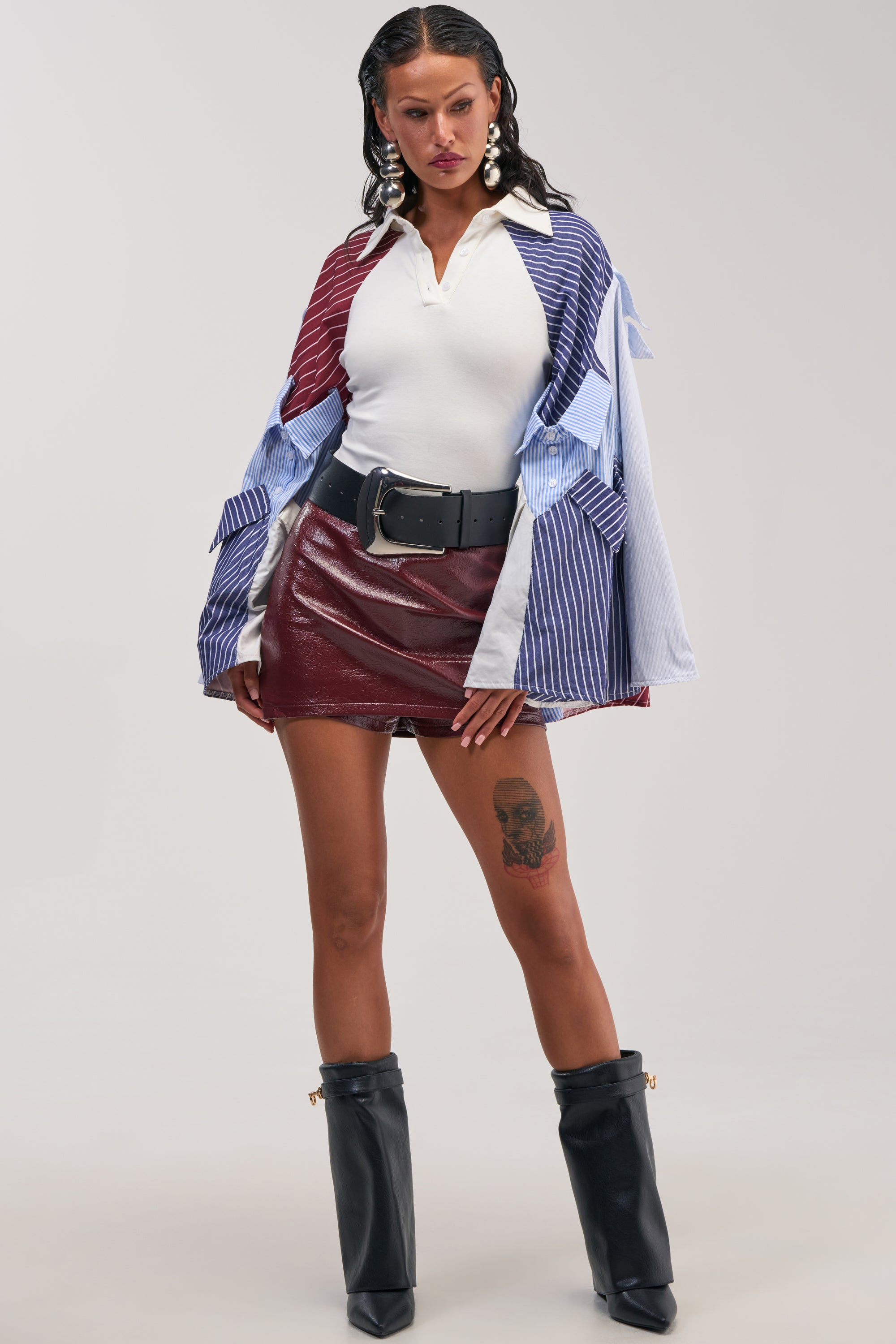 A woman poses confidently in the DOUBLE ZIP MINI SKIRT IN BURGUNDY, styled with a white polo shirt, color-blocked jacket, wide black belt, black boots, and large earrings. She has a thigh tattoo and wet-look hair.