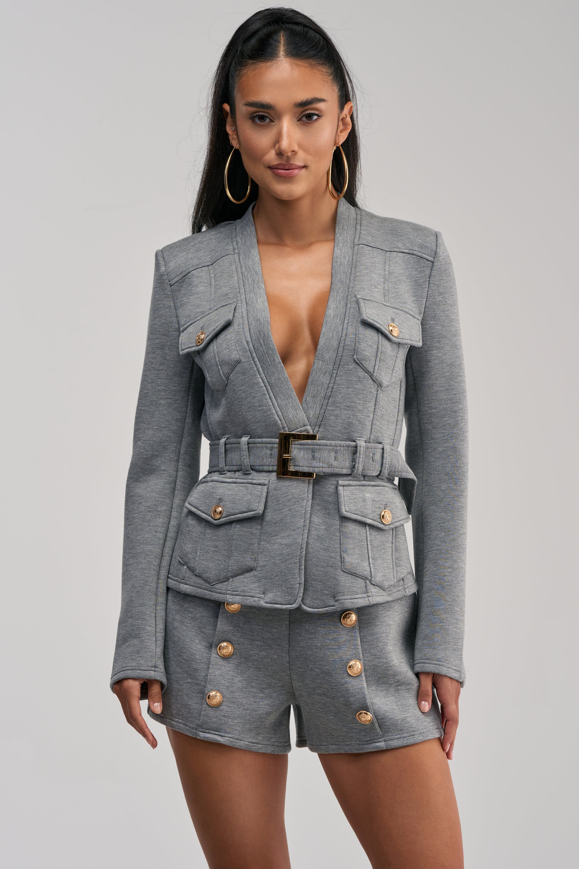 Model wears the LOLITA SCUBA BLAZER in gray, featuring gold buttons and a belted waist, paired with matching high-waisted shorts. She poses against a plain background with long dark hair, large hoop earrings, and a confident expression—perfectly capturing the office siren look.