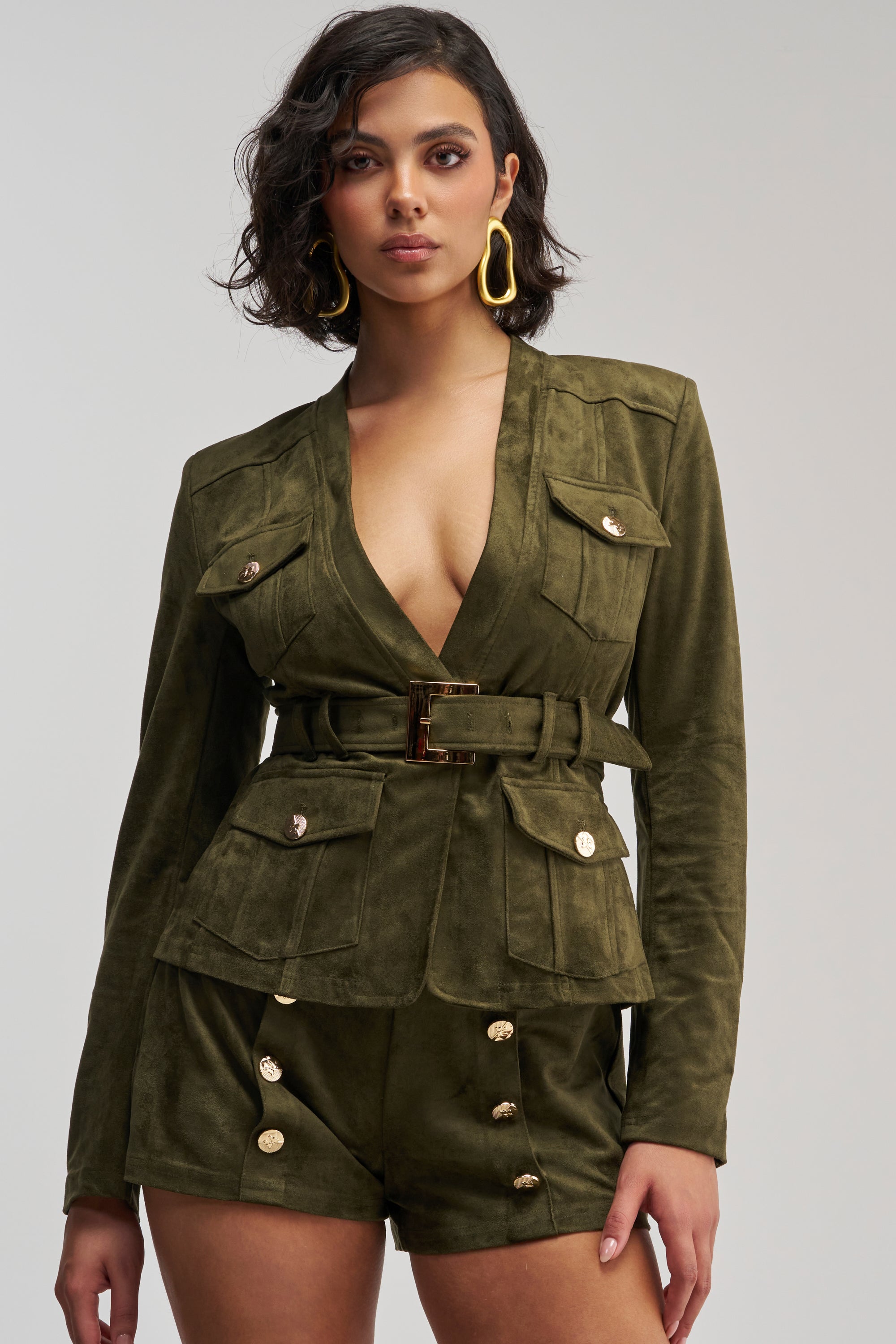 A woman stands facing forward against a plain background, wearing the LOLITA FAUX SUEDE BLAZER in olive green, styled with a belt and gold buttons, paired with matching high-waisted shorts and accessorized with large gold earrings.