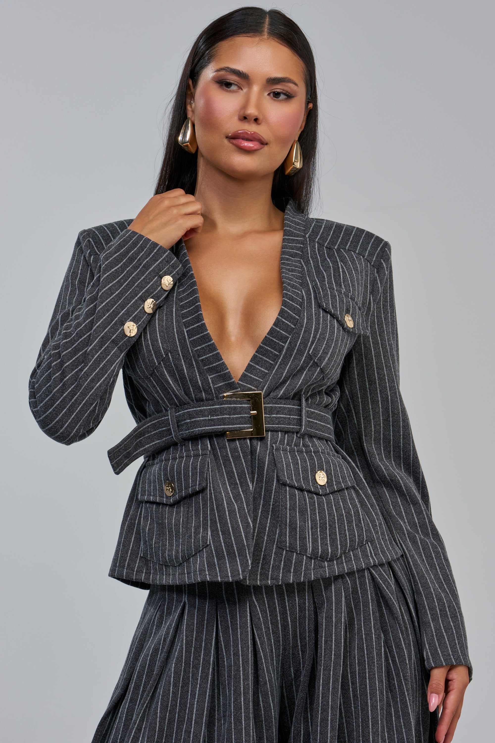 A woman wears the ROBYN PINSTRIPE BLAZER, a charcoal grey business casual piece featuring a deep V-neck and gold buttons, paired with matching wide-leg pants. She styles her long dark hair with gold earrings and poses confidently against a neutral backdrop.