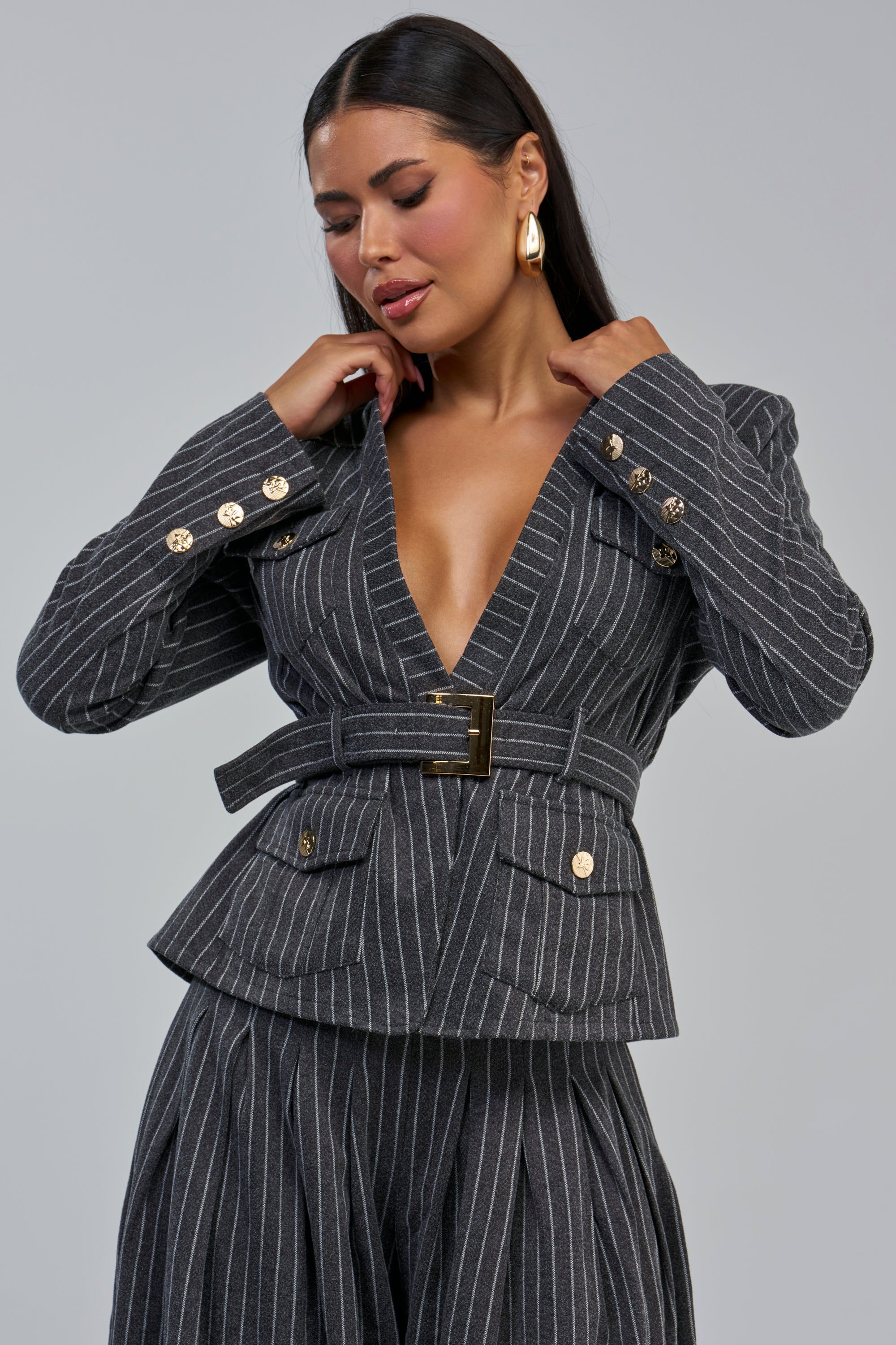Wearing the ROBYN PINSTRIPE BLAZER in charcoal grey with gold buttons and matching pants, she adjusts her collar while looking down. Long dark hair and gold hoop earrings complete her elevated business casual style.