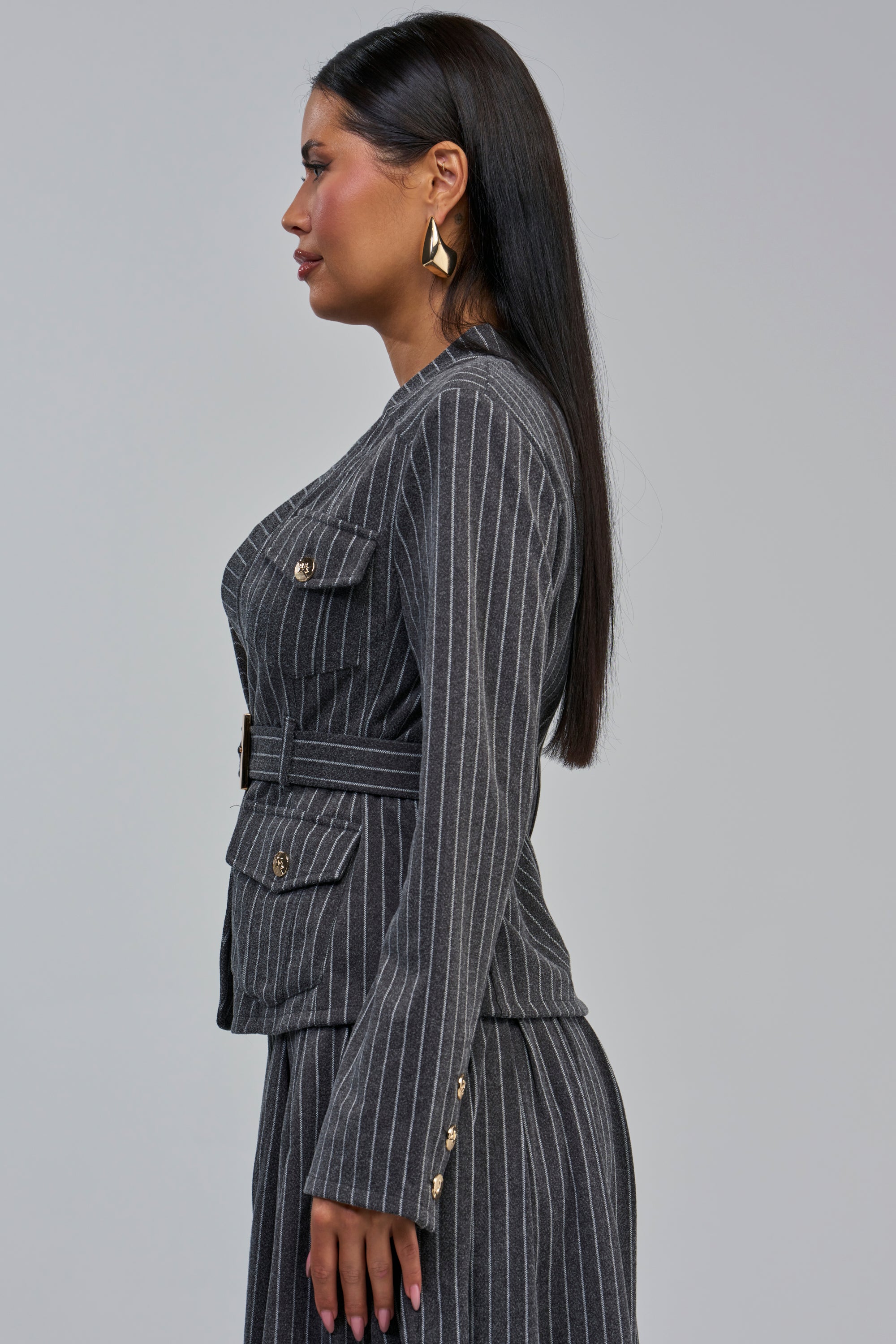 Wearing the ROBYN PINSTRIPE BLAZER in charcoal grey, this woman stands in profile against a plain light gray background. Her long straight dark hair falls past gold earrings, while gold buttons accent both her blazer and matching skirt.