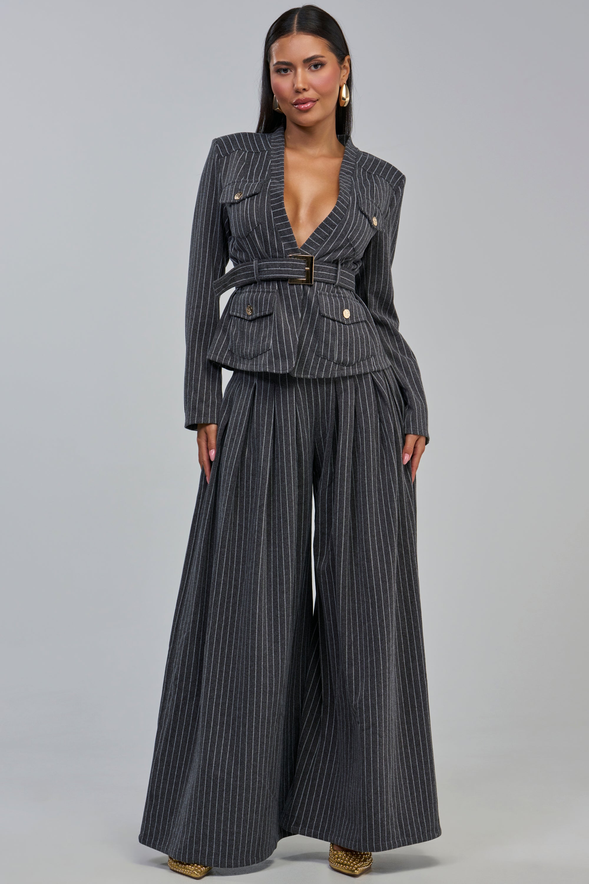 A woman models the ROBYN WIDE LEG PINSTRIPE TROUSER, paired with a matching belted deep V-neck blazer for a sophisticated, chic office look. She has long dark hair and gold earrings, standing against a plain light gray background.