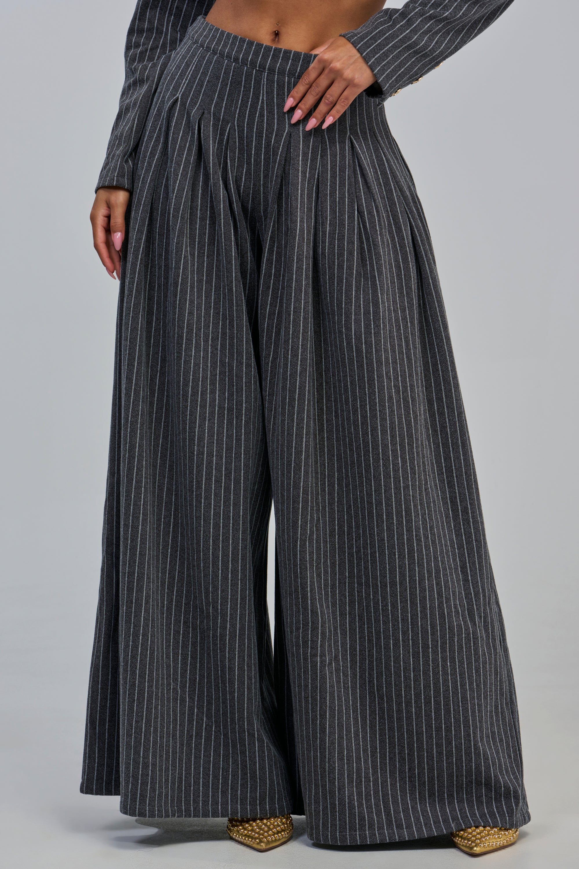 Wearing the ROBYN WIDE LEG PINSTRIPE TROUSER in a high-rise, wide-leg charcoal grey design with pinstripes and pleats—ideal for polished office looks—the individual poses with a hand on their hip and pointed gold shoes. The image is cropped just below the shoulders.