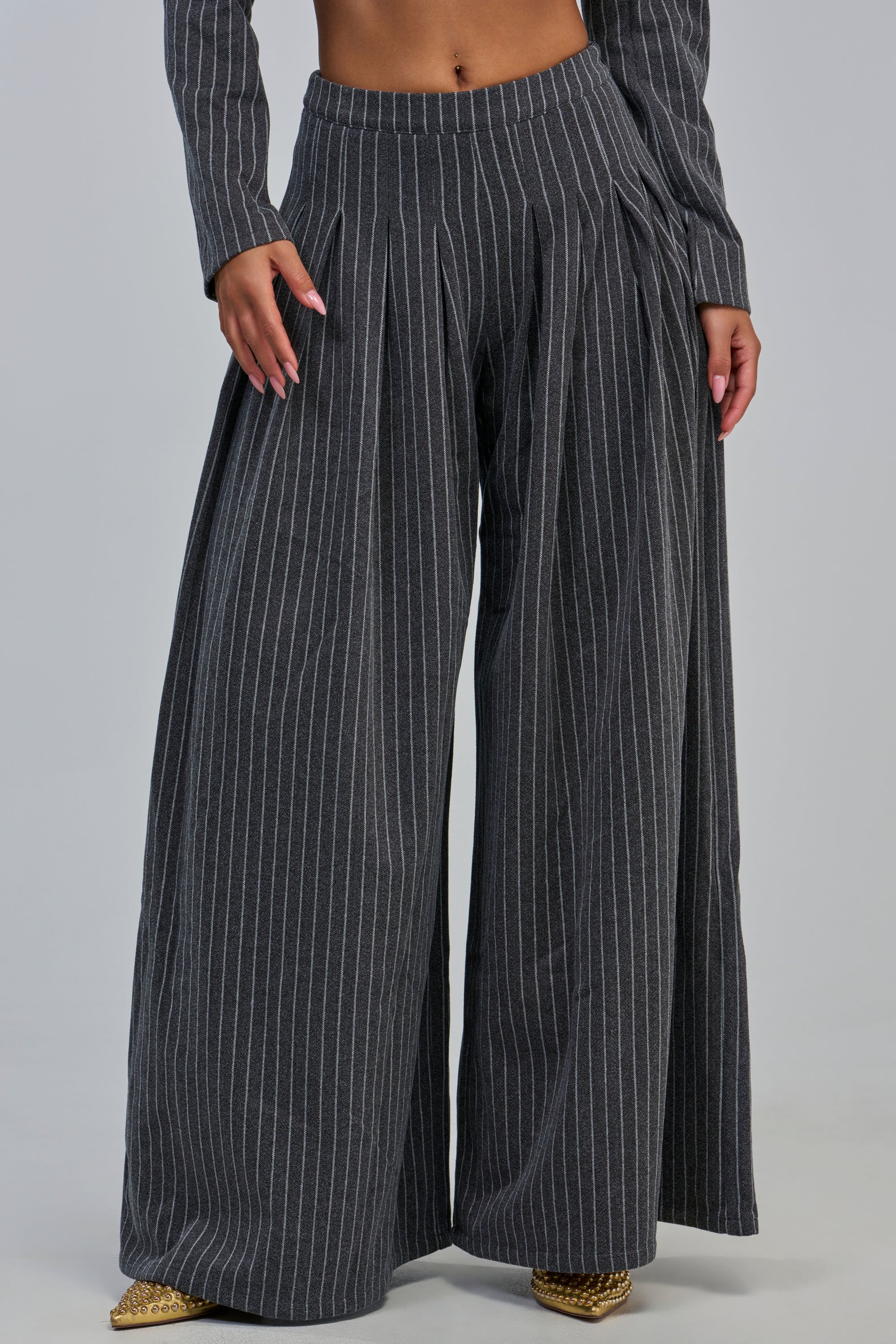 A person models the ROBYN WIDE LEG PINSTRIPE TROUSER in charcoal grey, featuring a high-rise, wide-leg silhouette with classic pinstripes. Visible from the waist down against a light gray background, the look offers a chic and modern approach to office attire.
