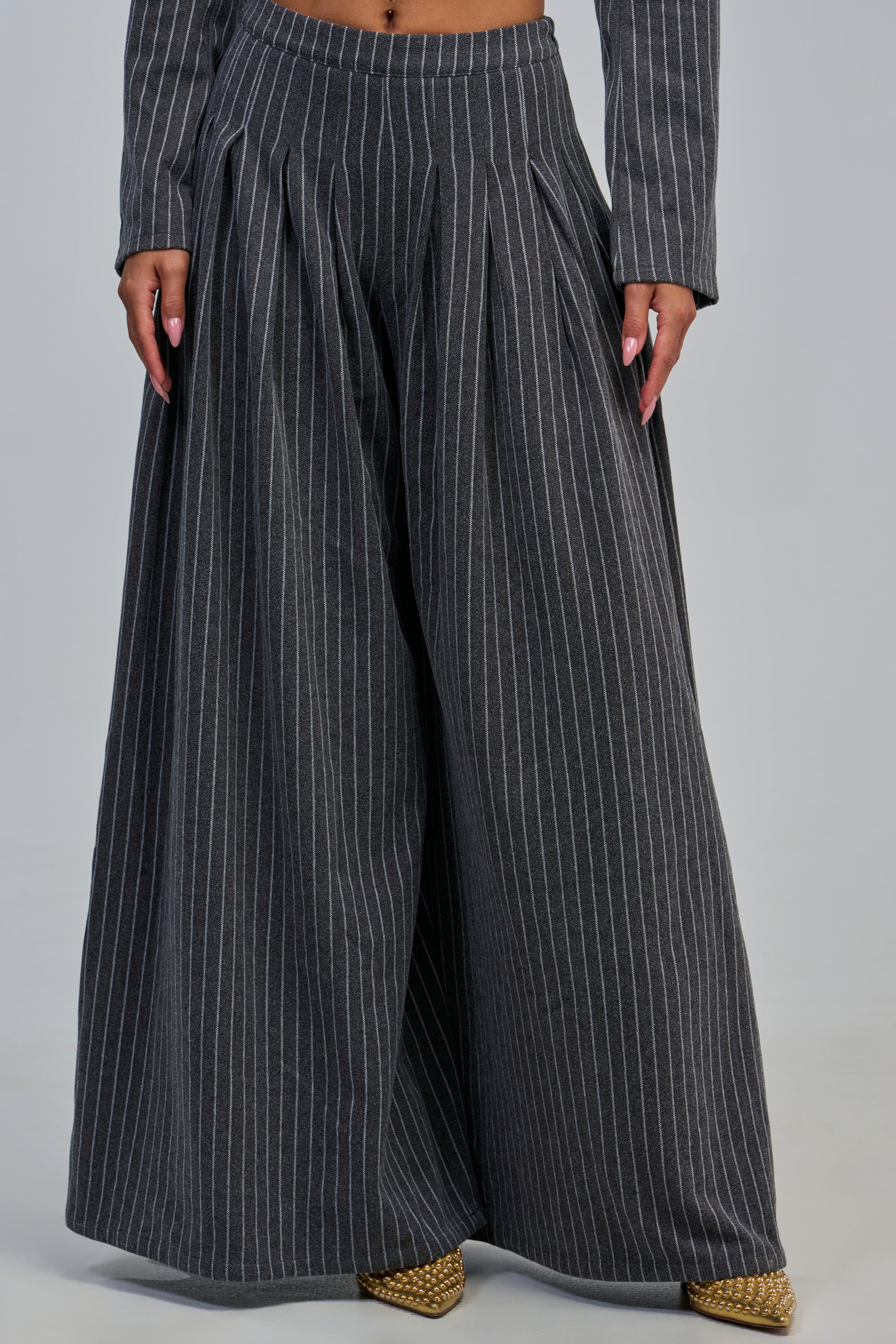 A person wears the ROBYN WIDE LEG PINSTRIPE TROUSER in a high-rise, wide-leg silhouette with charcoal grey pinstripes and pleats. The image shows only their lower torso, legs, and hands as they stand confidently in pointed, textured yellow shoes—an effortless fusion of modern office attire and bold style.