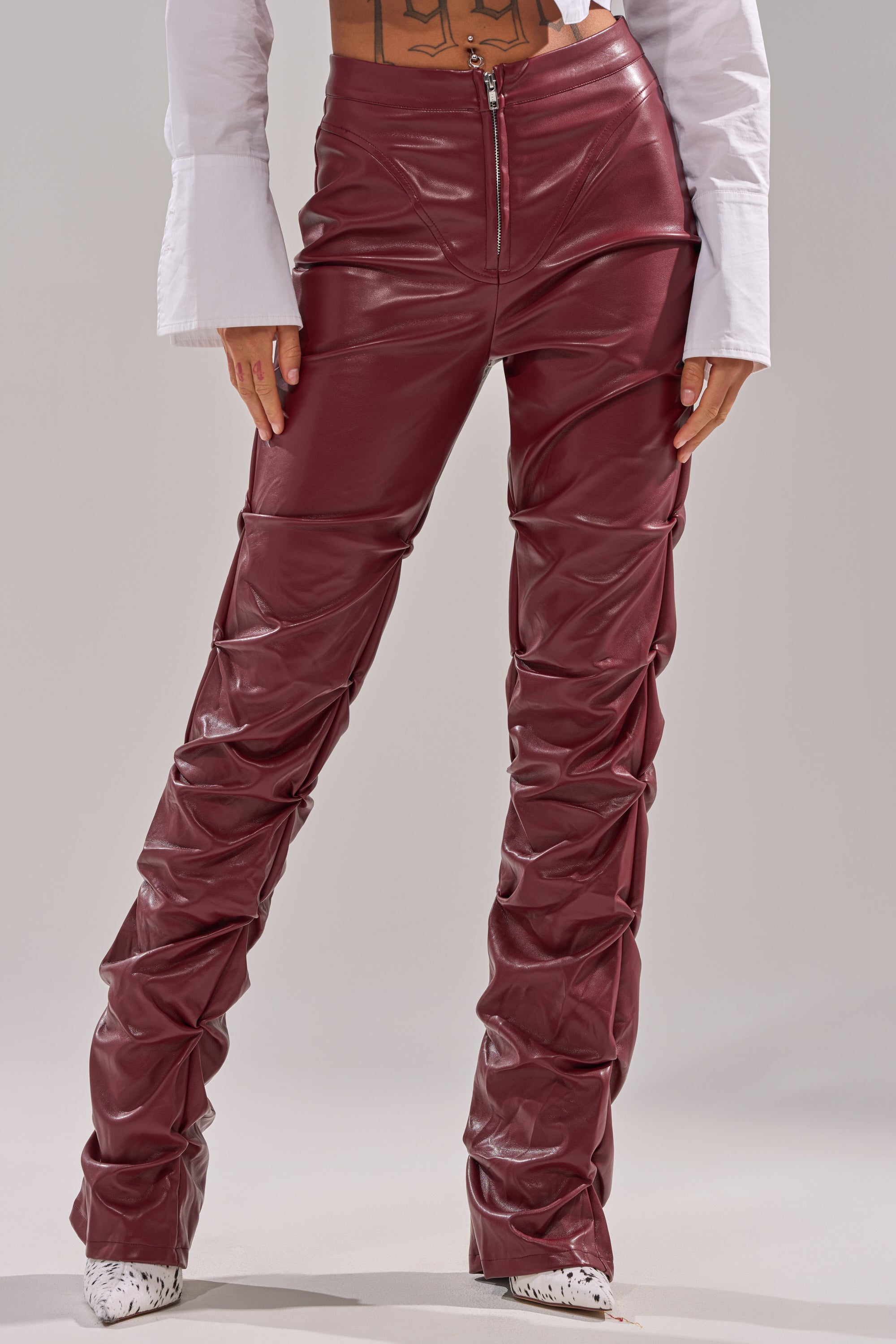 A person wears the DOWN AND DIRTY FAUX LEATHER PANT—shiny, ruched, burgundy faux leather with a mid-rise skinny fit—paired with white pointed-toe shoes and a white flared-sleeve shirt, against a light gray background. 0
