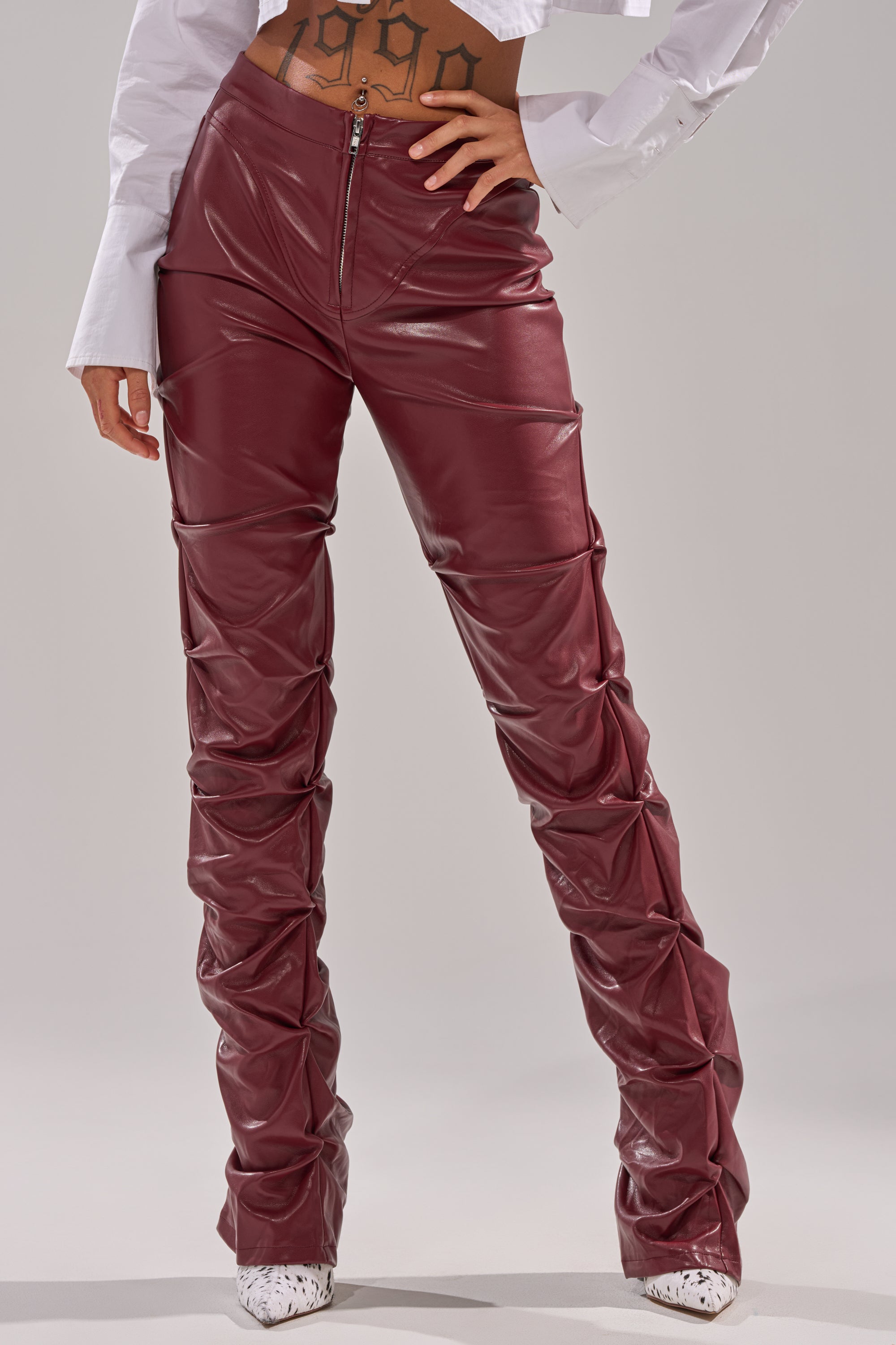 Someone wears the DOWN AND DIRTY FAUX LEATHER PANT—high-waisted, ruched burgundy faux leather pants—with a white cropped long-sleeve shirt and white pointed-toe shoes, showing their midriff and a stomach tattoo. 1