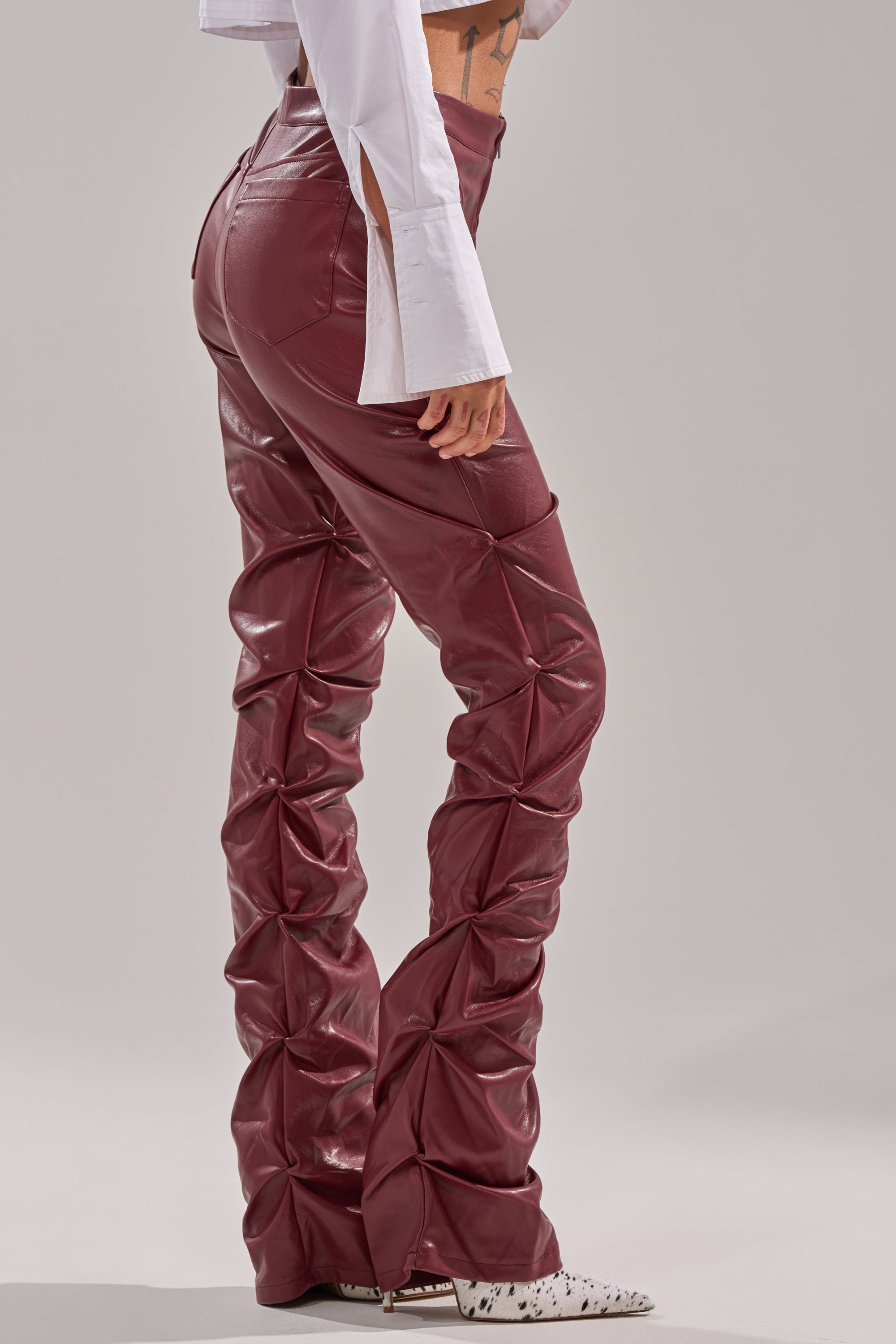 A person wears the DOWN AND DIRTY FAUX LEATHER PANT—a high-waisted, burgundy, skinny-leg pant with a ruched design—paired with a white long-sleeve top and speckled white shoes against a plain, light background. 3
